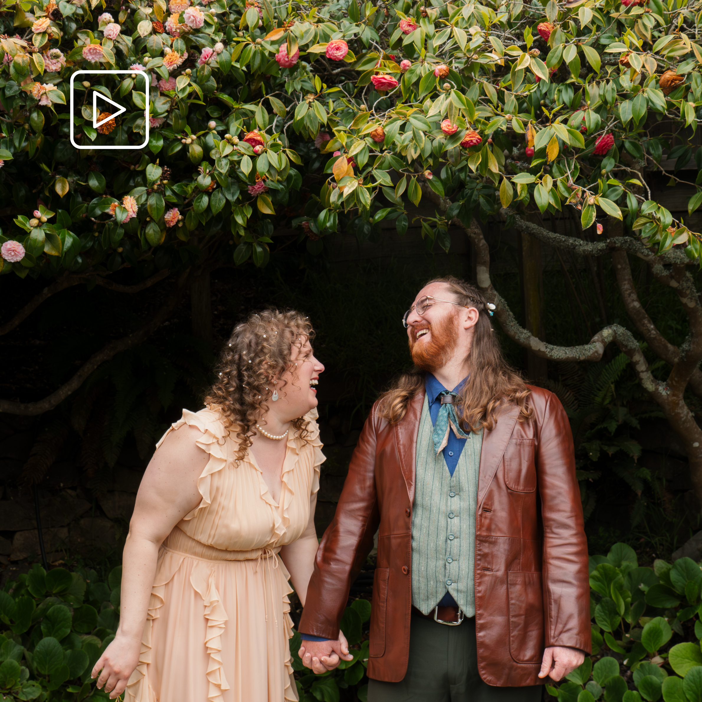 A smiling couple holding hands, standing outdoors in front of a large bush with pink and red flowers. The woman has curly blonde hair and is wearing a light peach dress, while the man has long hair, a beard, and is wearing glasses, a blue shirt, a brown leather jacket, and dark green pants.