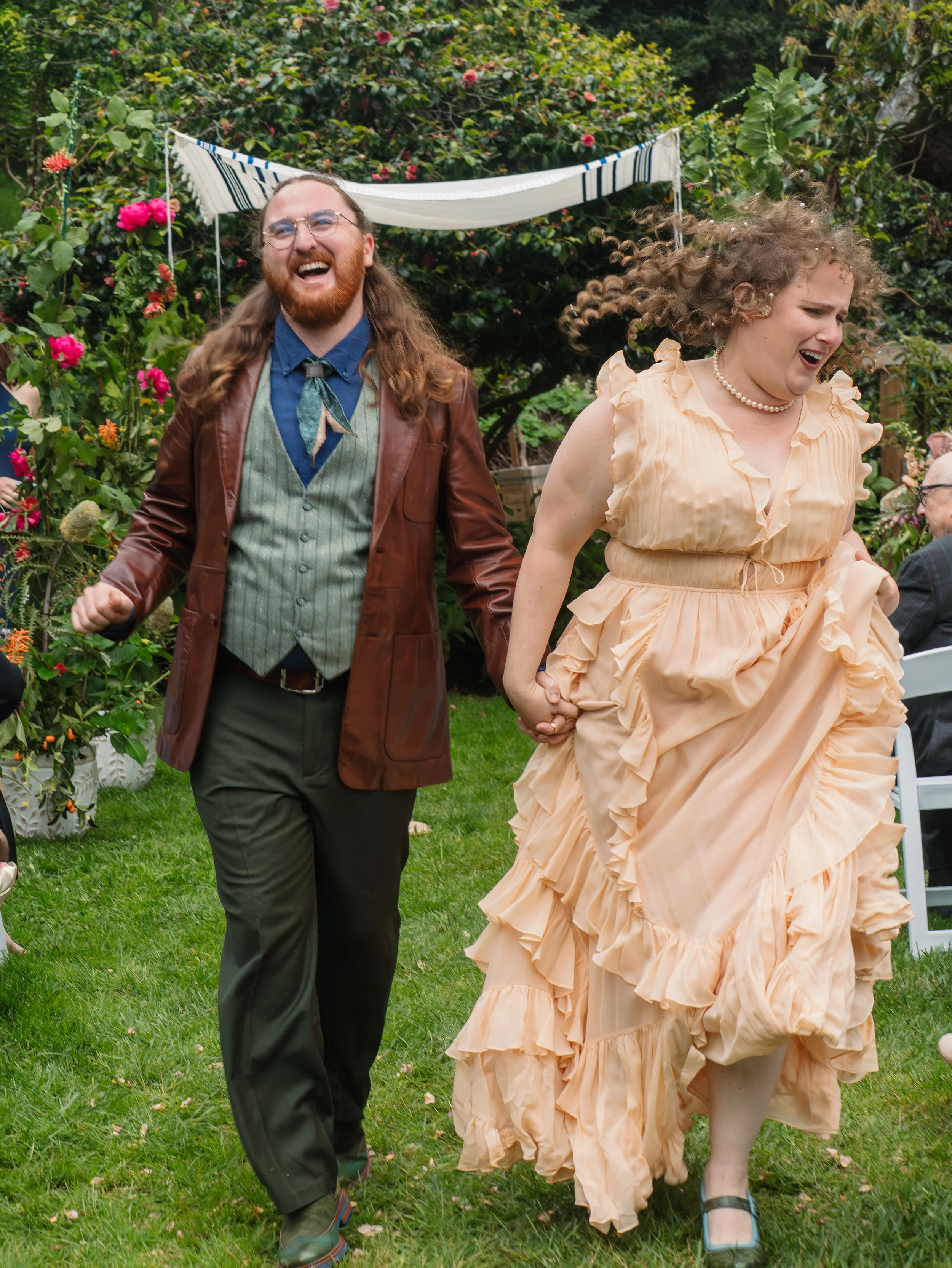 Two people are holding hands and running down the aisle in a garden, enjoying a joyful moment after their wedding ceremony, with greenery and pink flowers in the background.