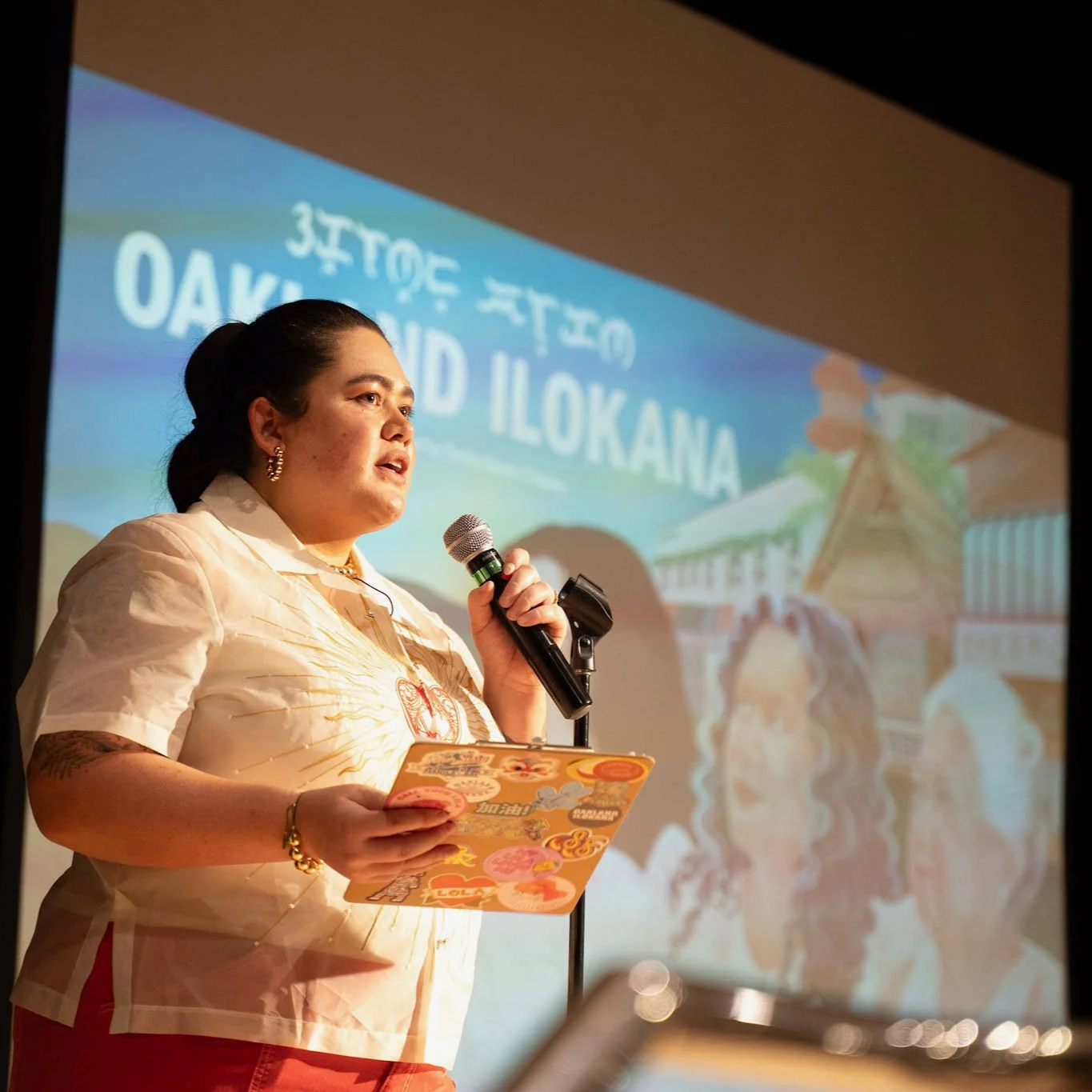 A woman holding a microphone and a colorful paper in front of a projected presentation slide with the words 'OAKLAND ILOKANA' and an illustration of a traditional building.