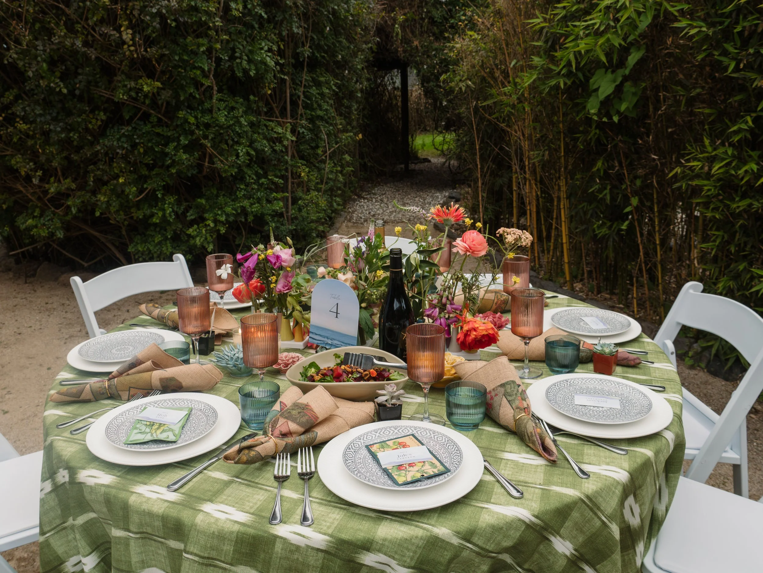 A round outdoor table set for a meal with a green plaid tablecloth, surrounded by white chairs. The table features colorful flowers, glasses, plates, utensils, and small menu cards. The background shows dense green shrubbery and a pathway with a gate