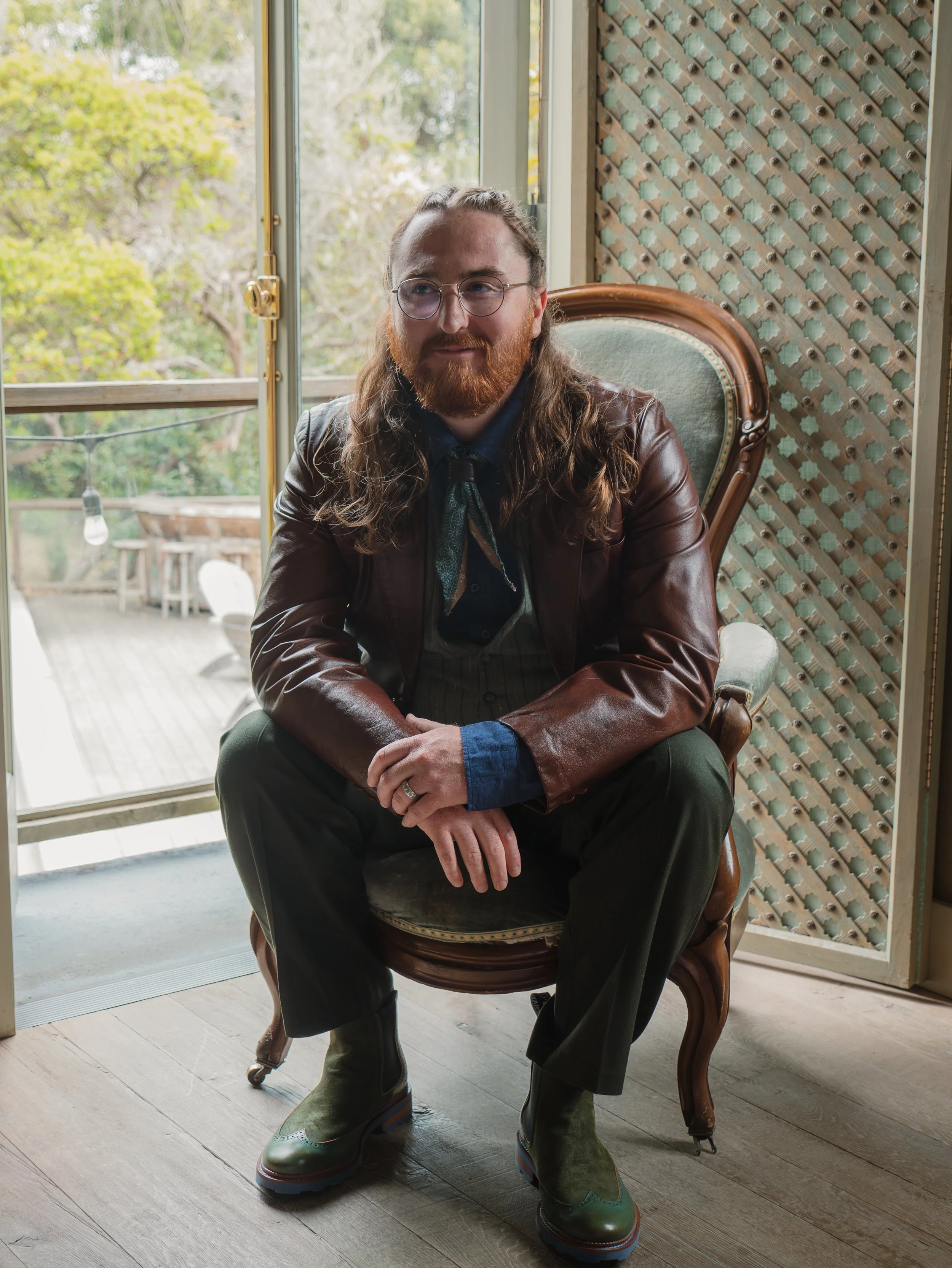 A man with long hair, glasses, and a beard sitting in an antique wooden chair. He is wearing a dark shirt, a green tie, a brown leather jacket, and green boots. He is indoors near a large window with a blurred outdoor view of trees and a deck area.