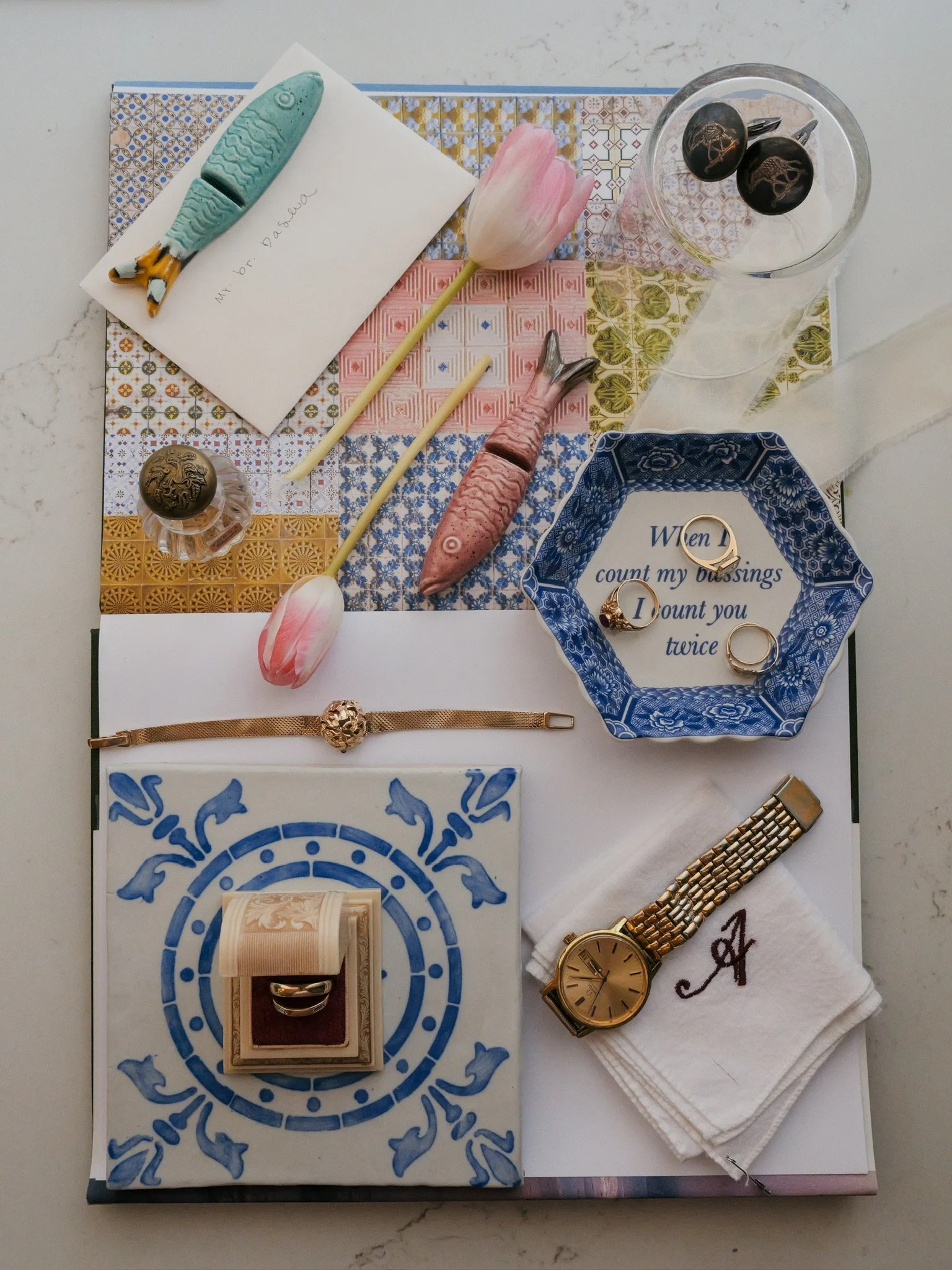 Flat lay of various personal items on a patterned blue and white cloth, including a gold wristwatch, a gold ring, a pair of black and gold cufflinks, a pink and white tulip, a ceramic fish-shaped pen, a white towel with text, and a blue decorative bo