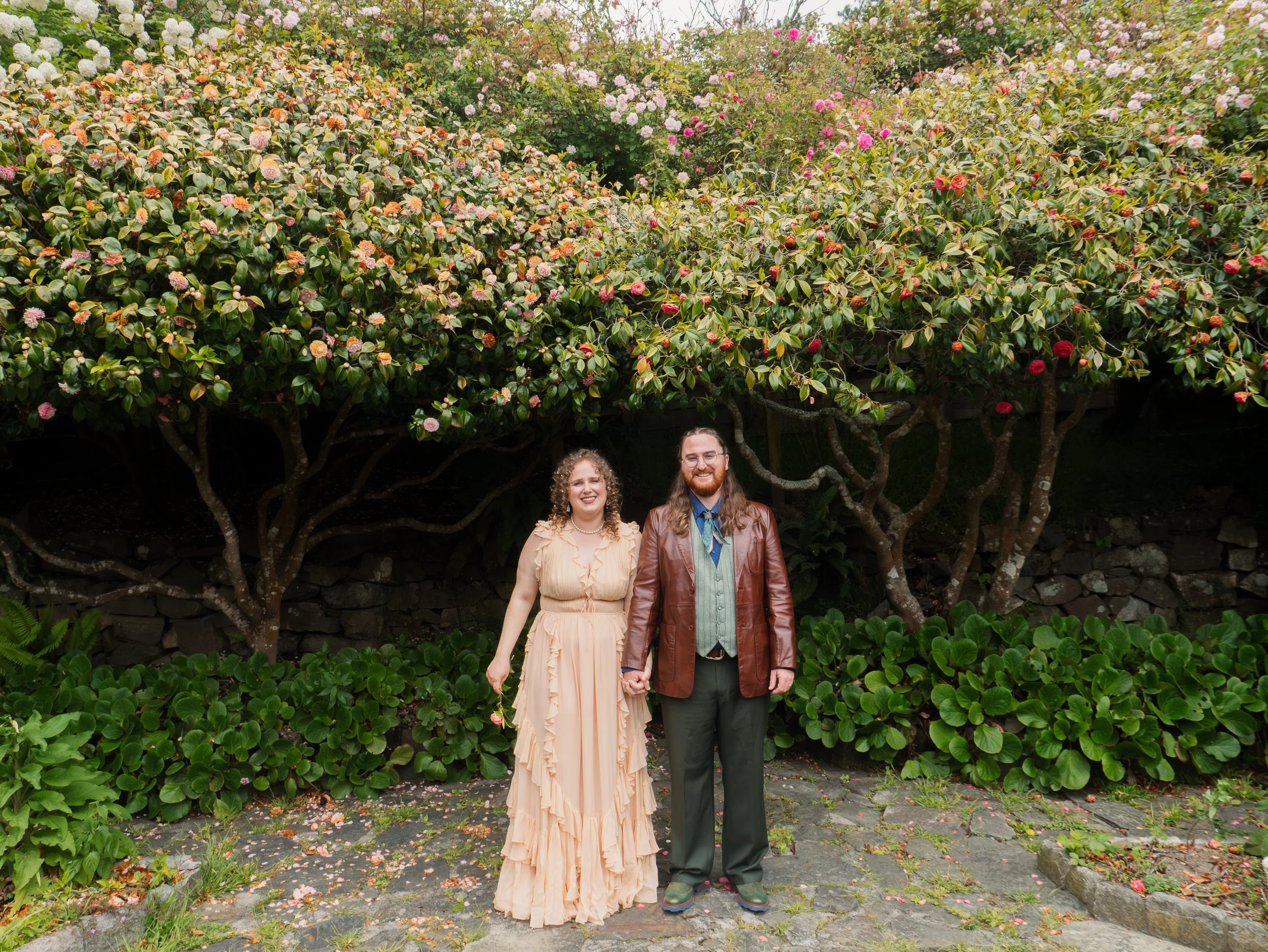 A couple holding hands standing under blooming pink and white roses in a garden.