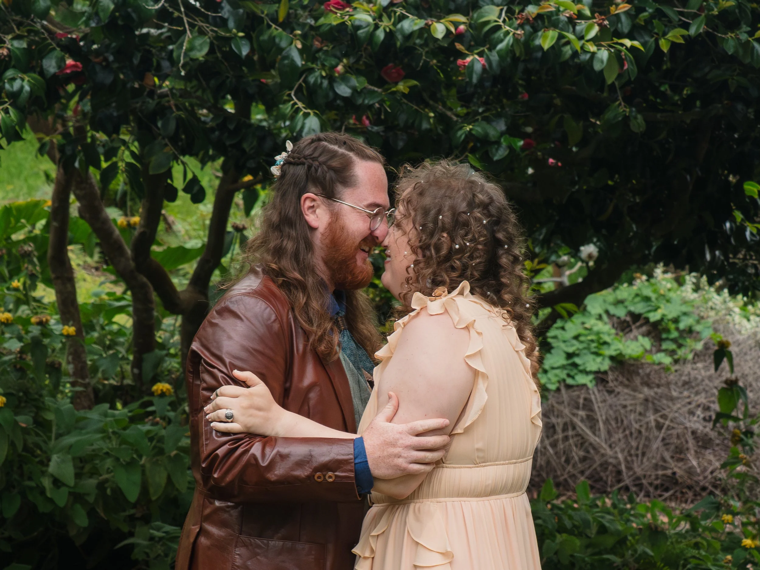 A man and woman standing close, smiling and touching foreheads, in front of a lush green hedge with pink flowers.