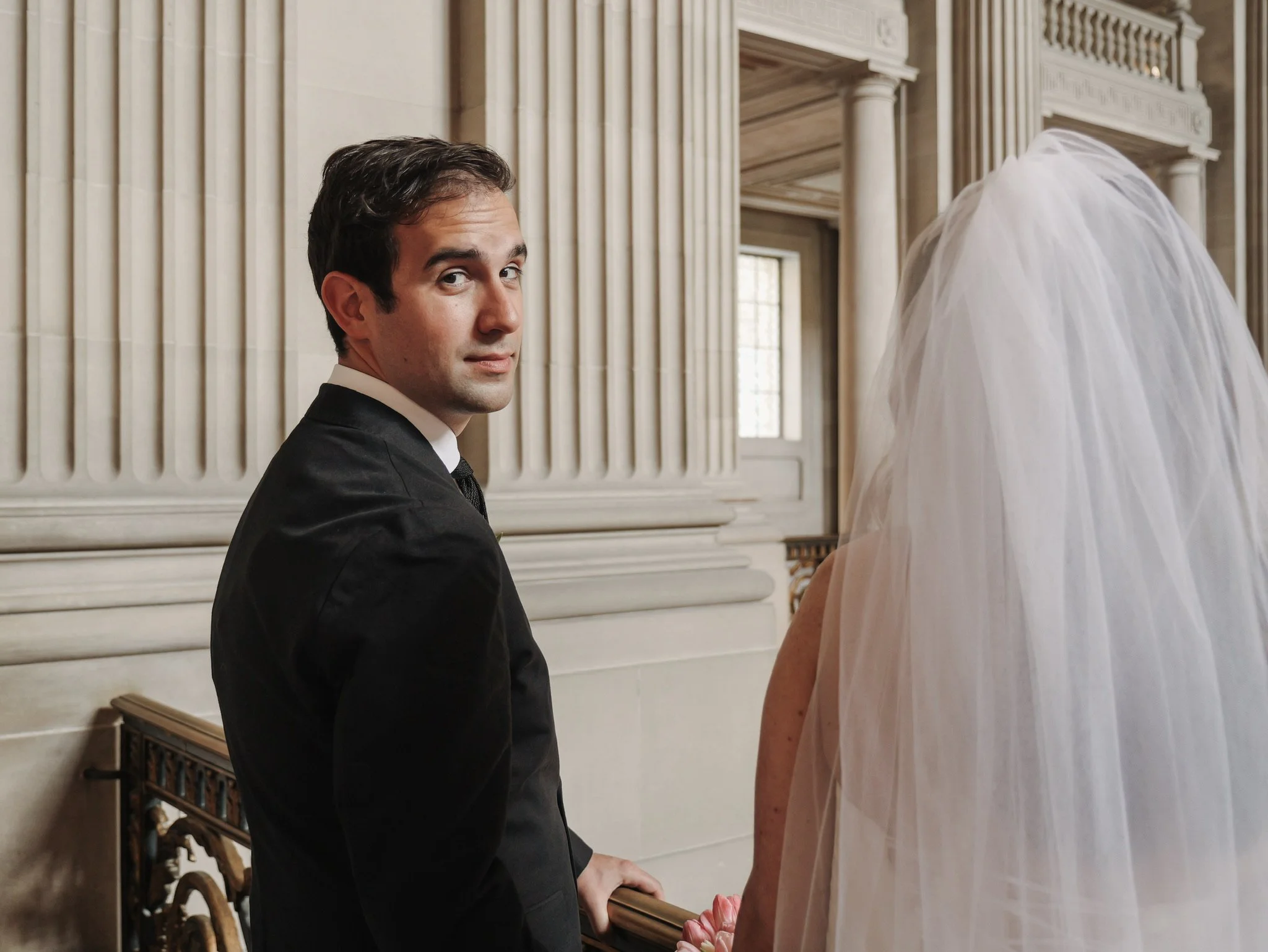 A man dressed in a black suit sitting on a bench inside San Francisco City Hall with tall columns and large windows, looking over his shoulder.