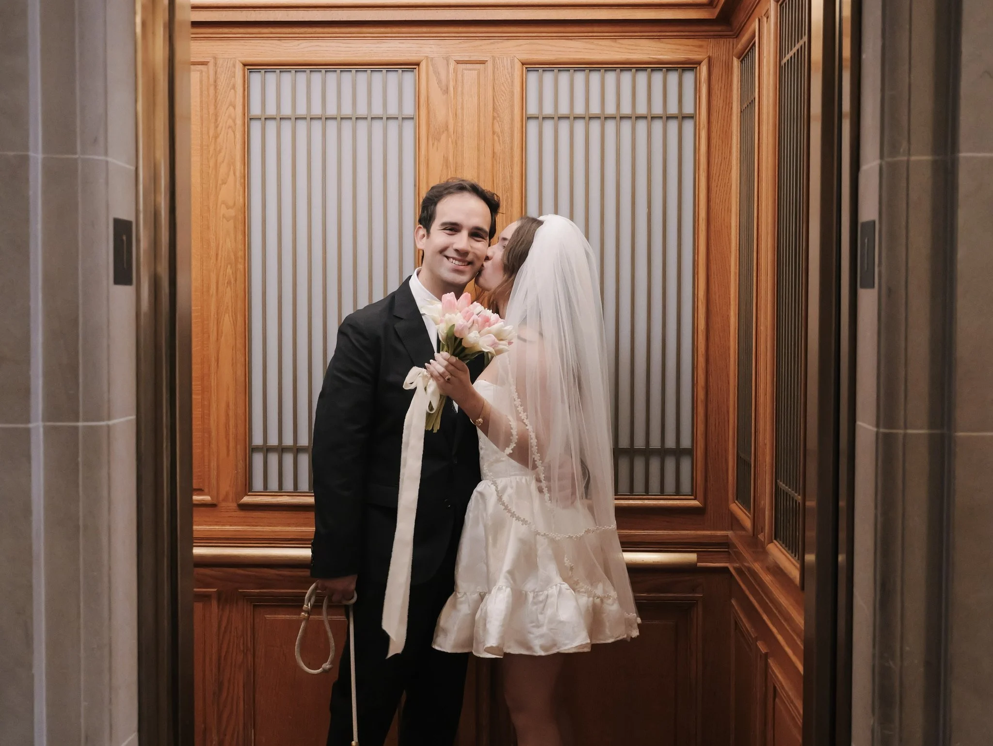 A newlywed couple in an elevator, with the bride planting a kiss on the groom's cheek. The groom is smiling and holding a bouquet of pink and white flowers, wearing a black tuxedo and white shirt. The bride is wearing a white wedding dress with a vei