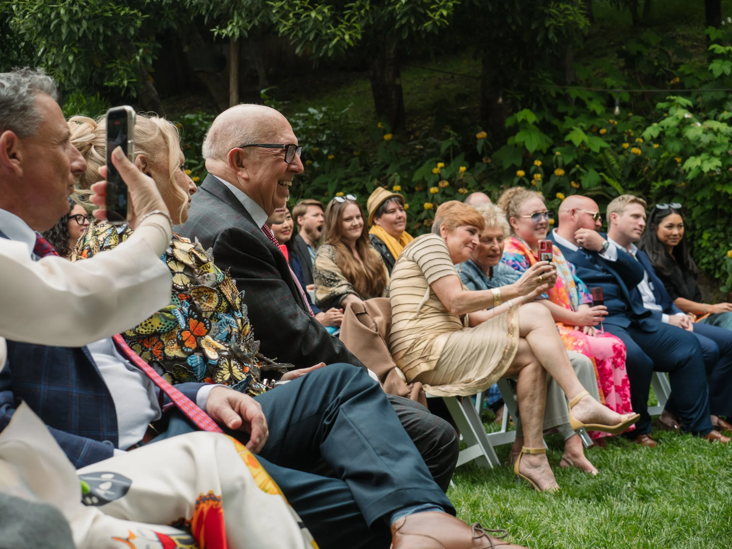 Group of people sitting outdoors smiling and watching an event, with lush green trees and foliage in background.