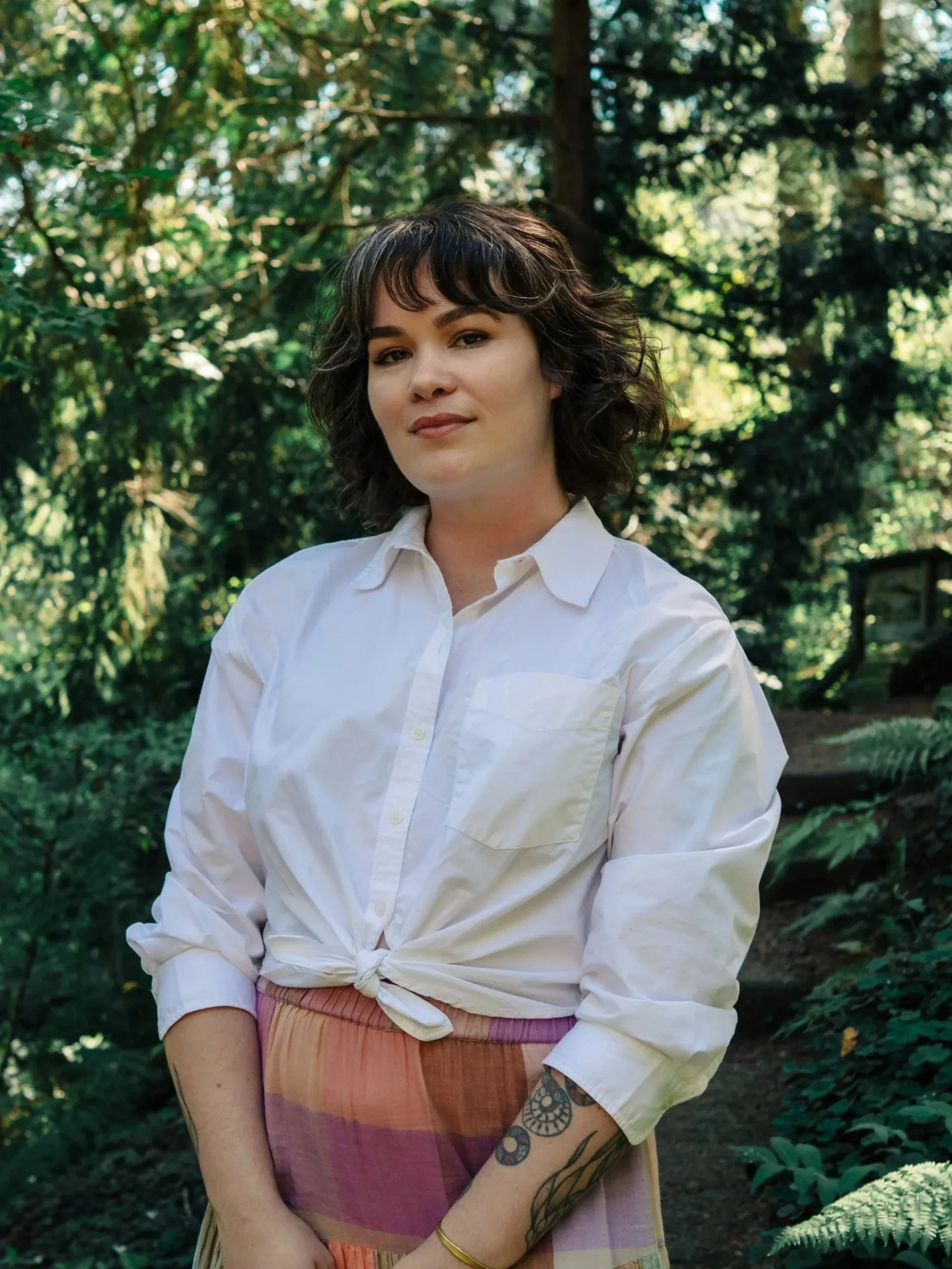 A woman with medium-length dark hair and tattoos on her left arm stands outdoors in front of green foliage, wearing a white button-up shirt tied at the waist and a multicolored skirt.