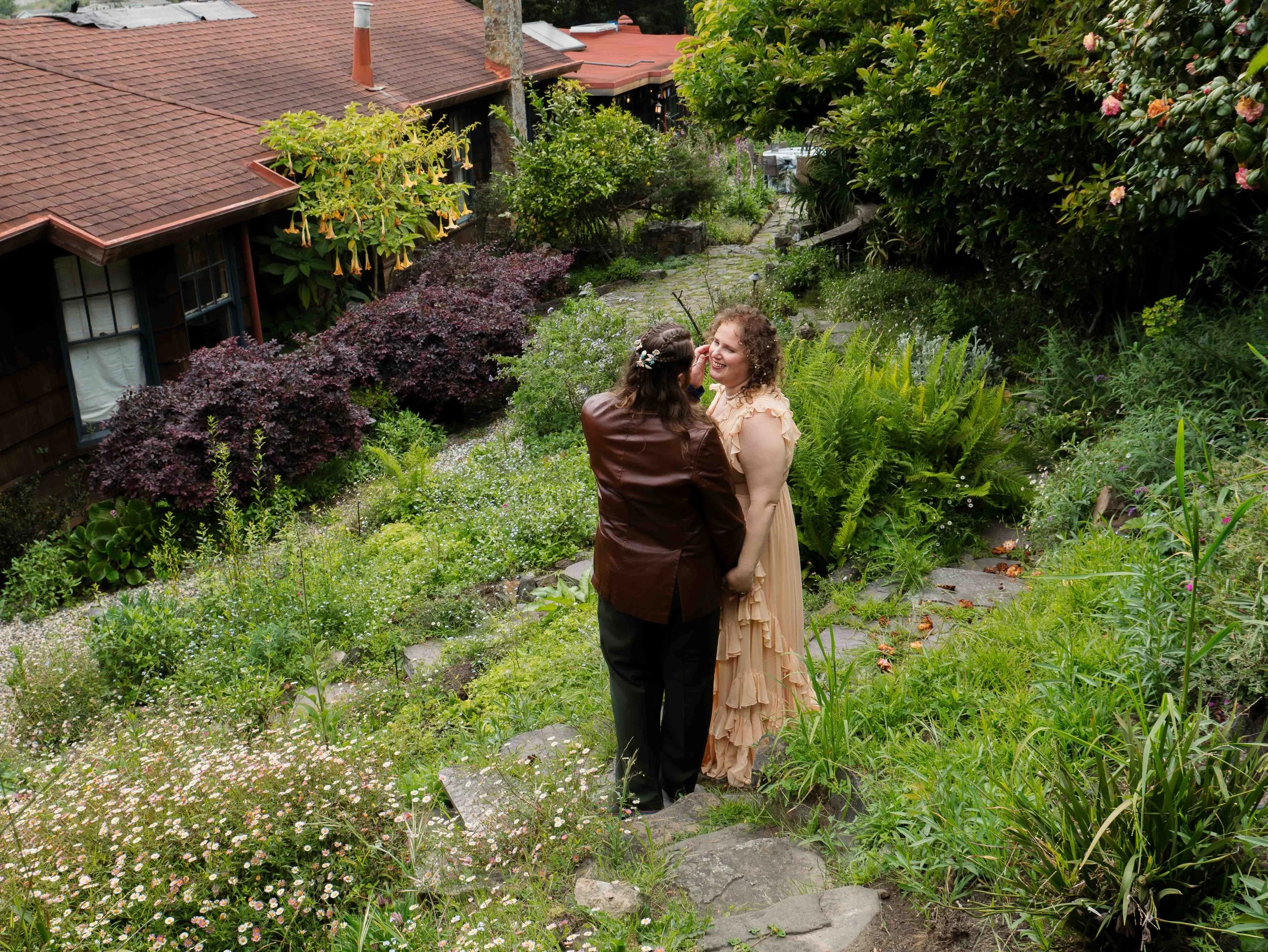 A couple smiling and interacting in a lush, green garden with a stone pathway, surrounded by flowering bushes and trees, with a house roof visible in the background.