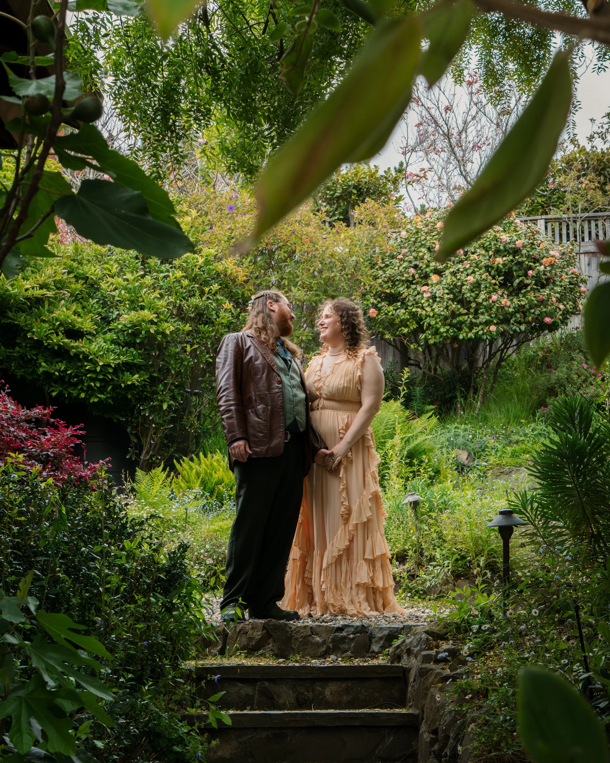 A couple standing close together in a lush garden surrounded by green plants and colorful flowers, looking at each other and smiling.