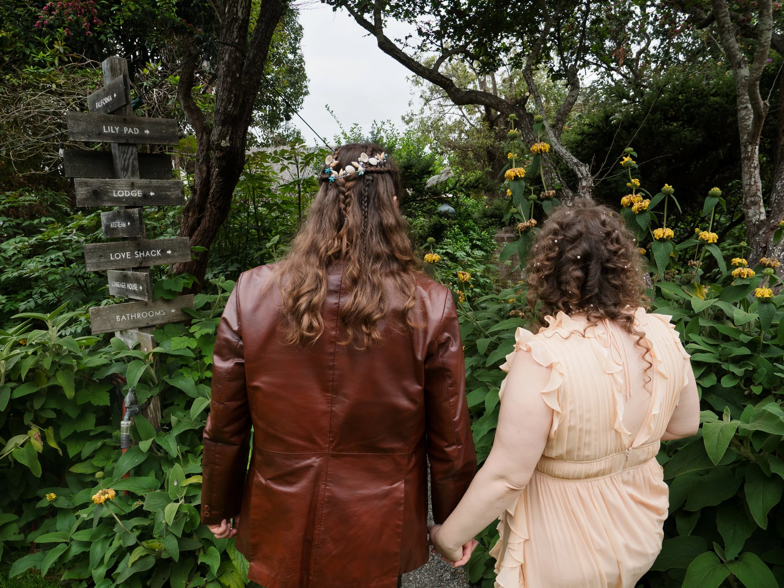 A couple holding hands walking through a lush garden with trees, flowers, and a wooden signpost with multiple directional signs.