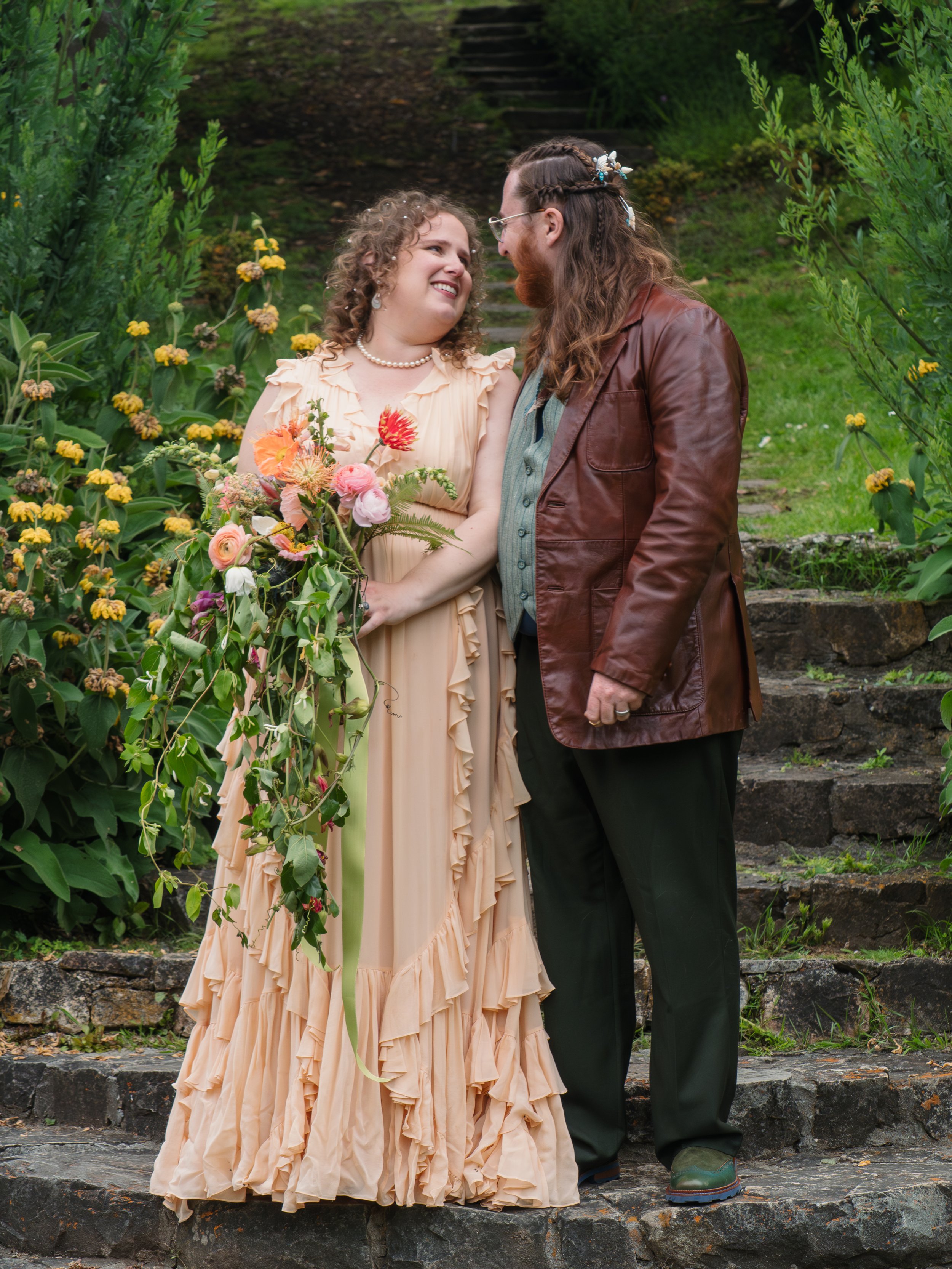 A couple standing on stone steps outdoors, close to each other, looking into each other's eyes, surrounded by lush green plants and flowers.