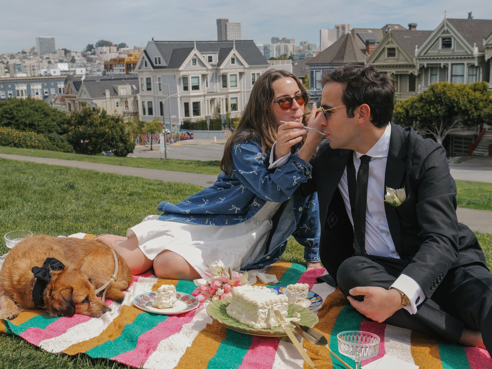 A couple having a romantic moment during a daytime outdoor celebration on a colorful blanket, with a picnic spread including cake, flowers, and drinks, in front of Victorian-style houses and a cityscape.
