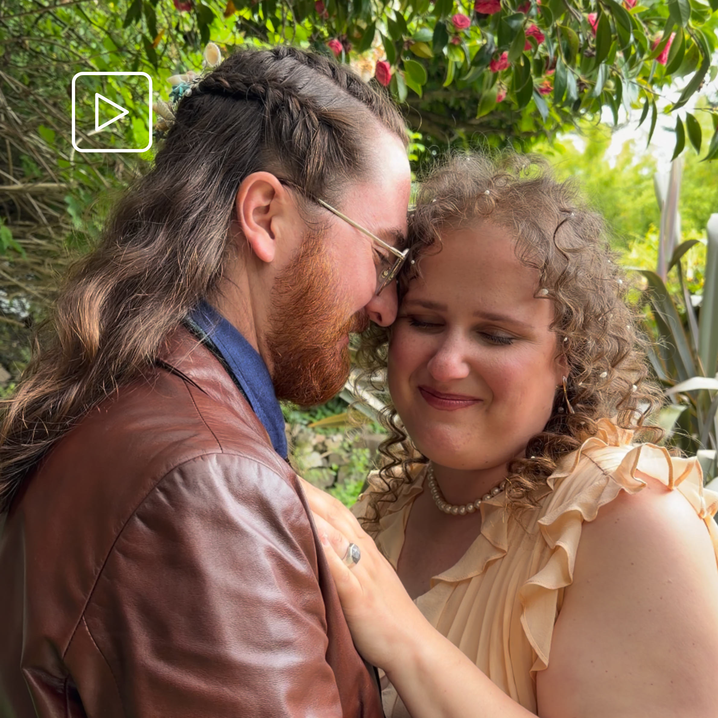 Close-up of two people in an intimate moment, touching foreheads. One has long, wavy hair and the other has curly hair with pearls in it. They are outdoors, with lush greenery and pink flowers in the background.