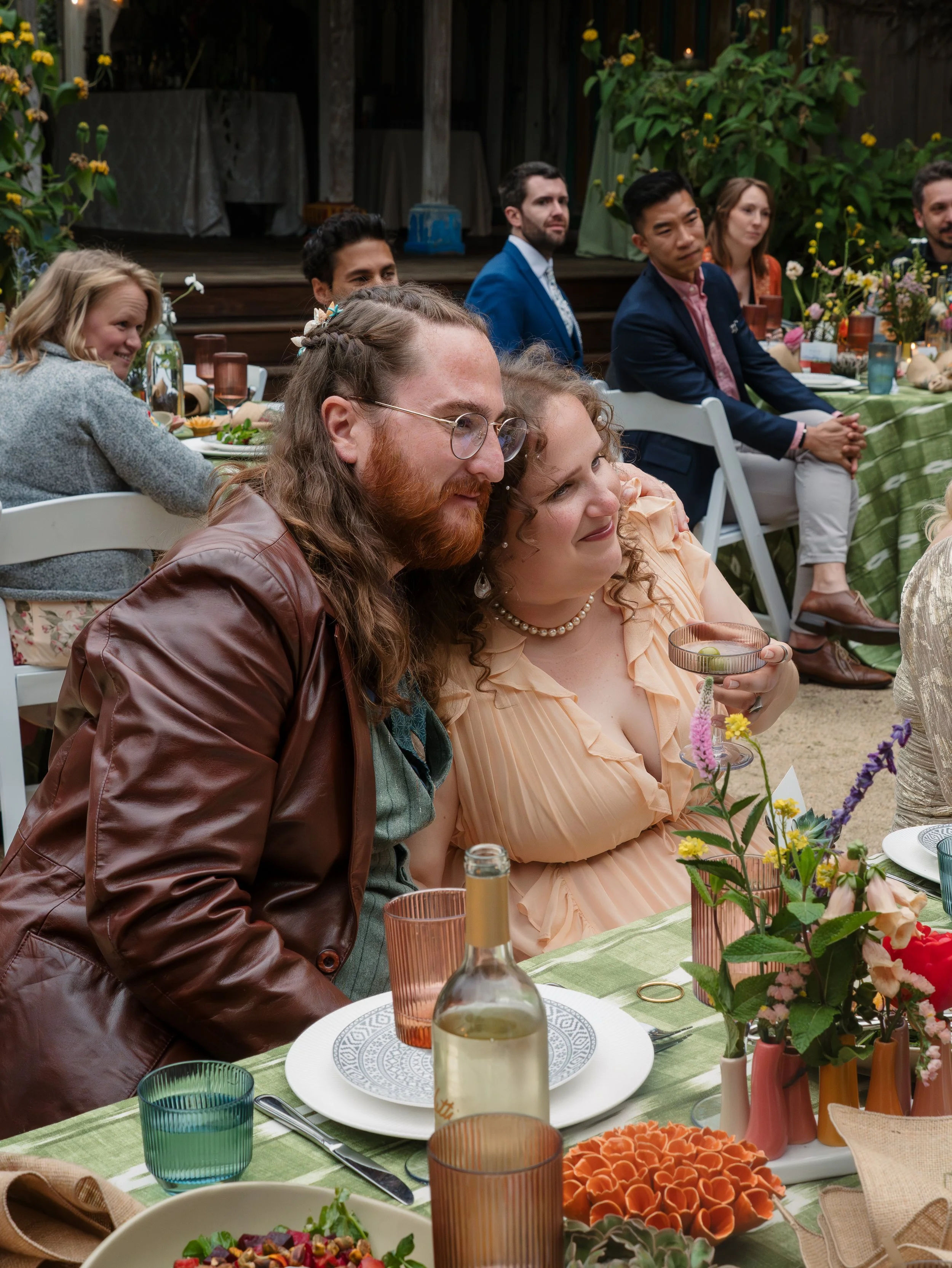People sitting at a long outdoor table decorated with flowers, with plates, glasses, and bottles, during daytime.