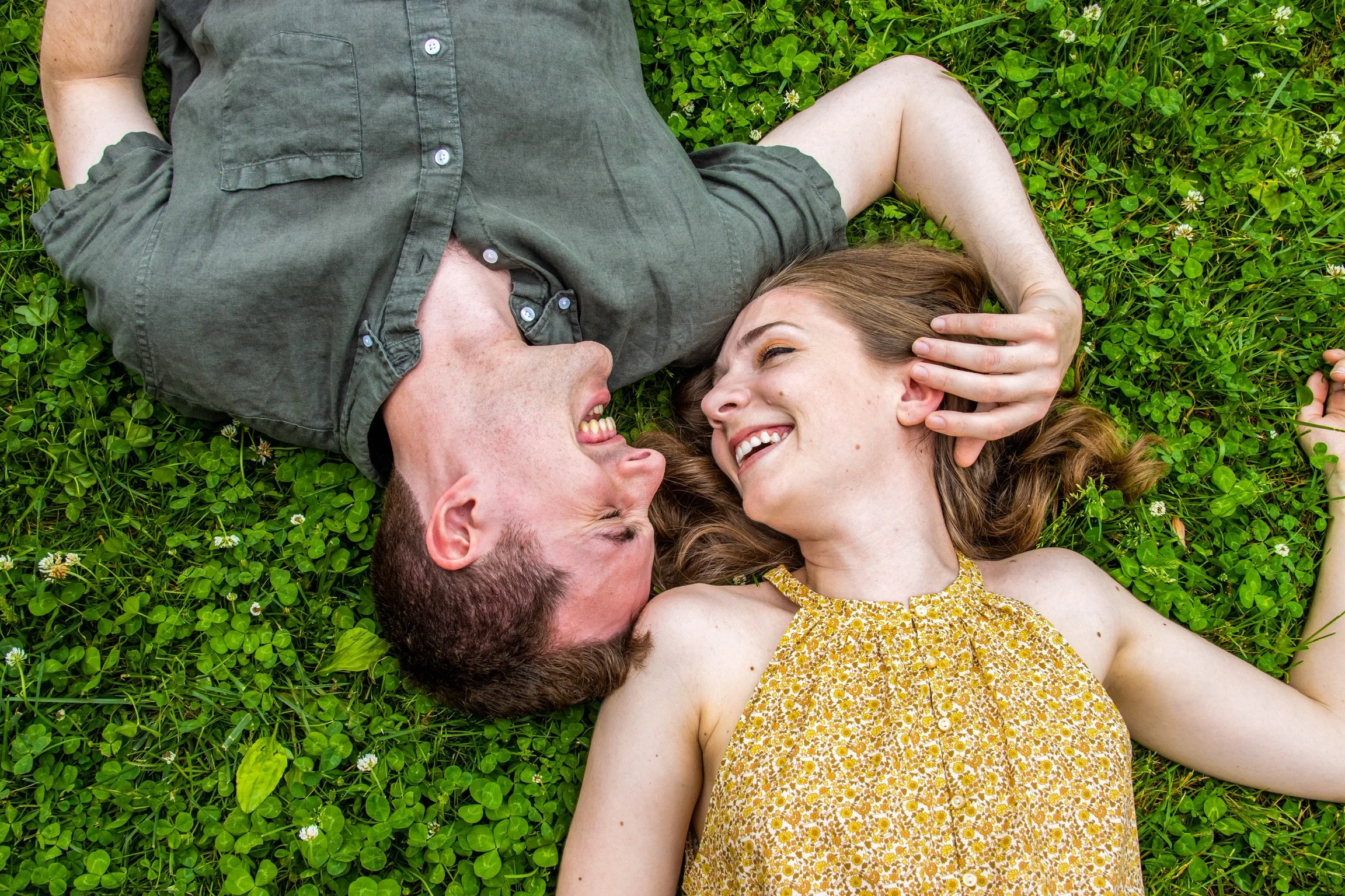 A man and woman lying on green grass, facing each other, smiling and touching each other's faces.