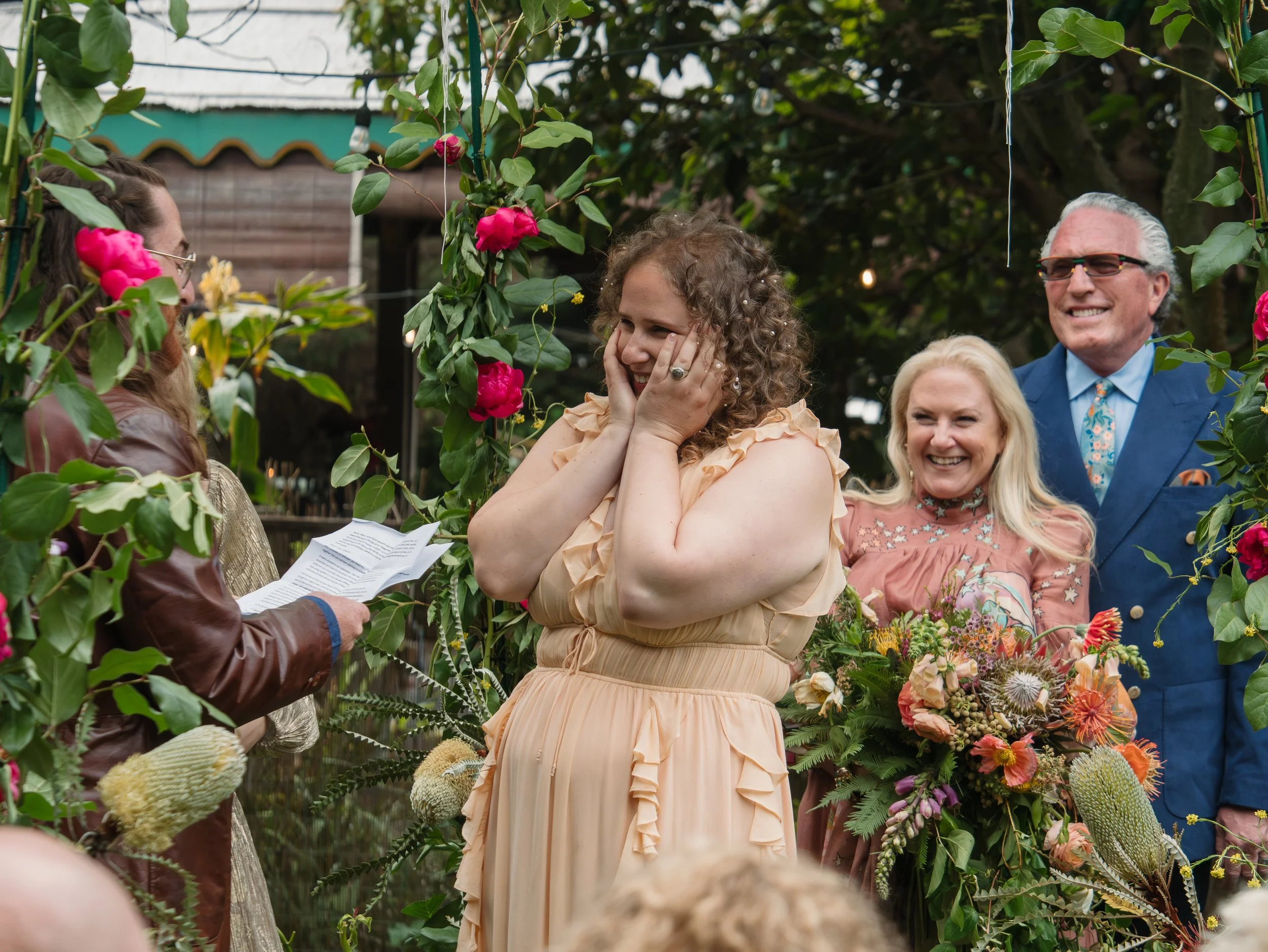 A woman in a peach-colored dress with curly hair and pearl hair accessories is smiling while holding her face with both hands during a celebration. She is surrounded by a group of smiling people, a man in a blue suit with sunglasses, a woman with blo