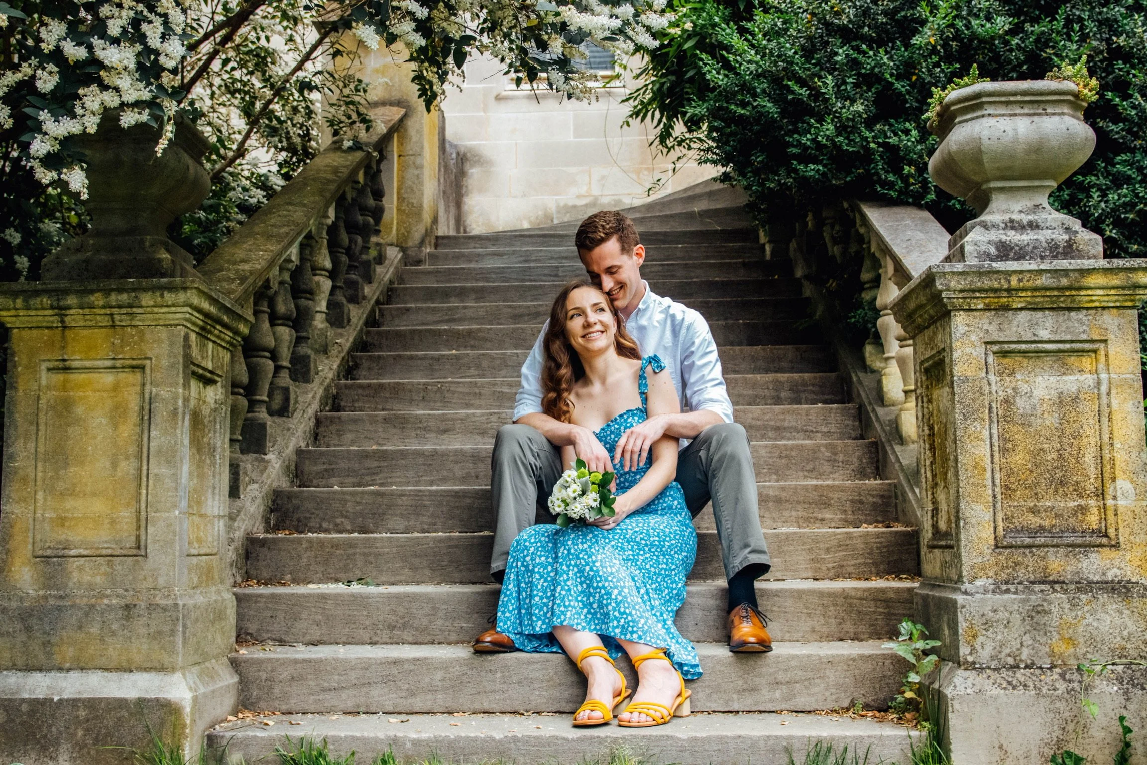 A happy couple sitting on outdoor stone stairs, surrounded by greenery and flowers, with the woman holding a bouquet of white flowers.