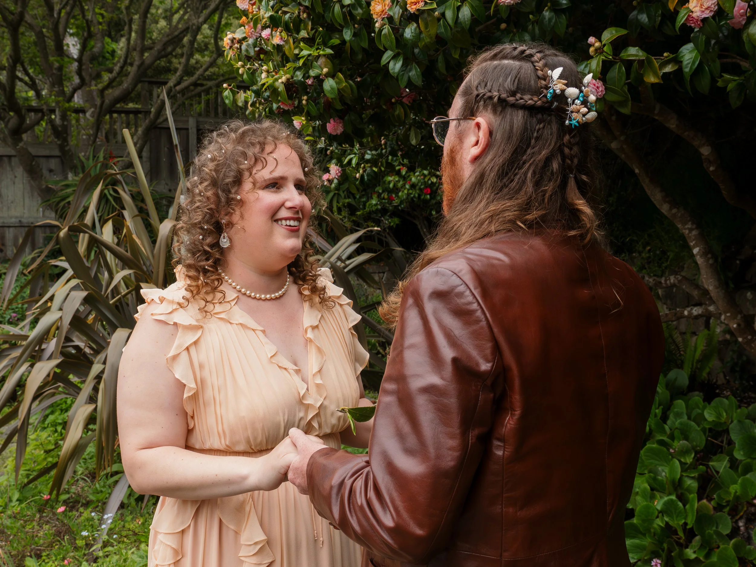 A man with long hair and glasses holding hands and smiling at a woman with curly hair wearing a peach ruffled dress with pearls and earrings, in a garden with flowering bushes and trees.