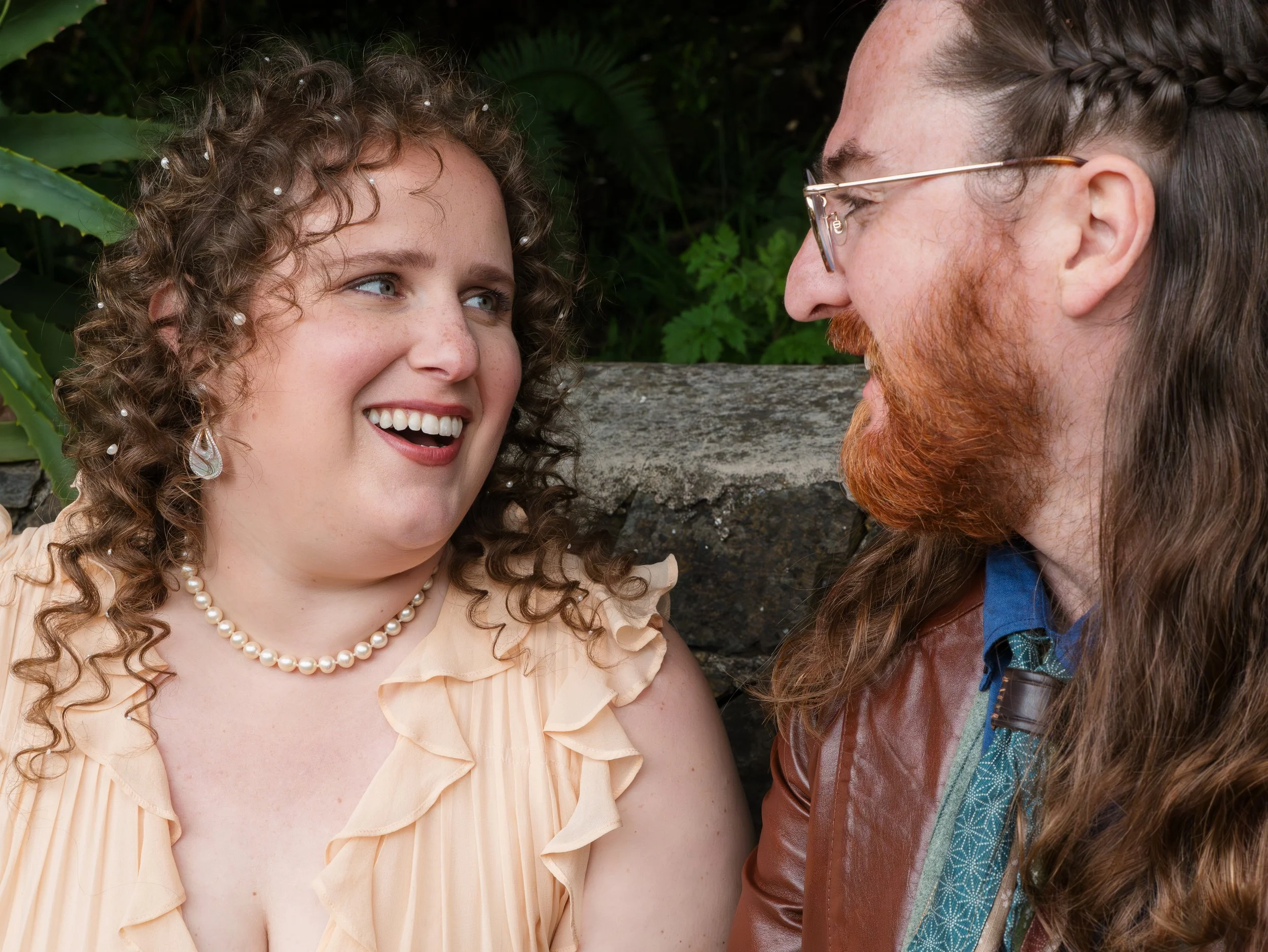 Close-up of a smiling woman with curly hair, pearls, and earrings speaking to a man with long hair, glasses, and a beard, outdoors with greenery.