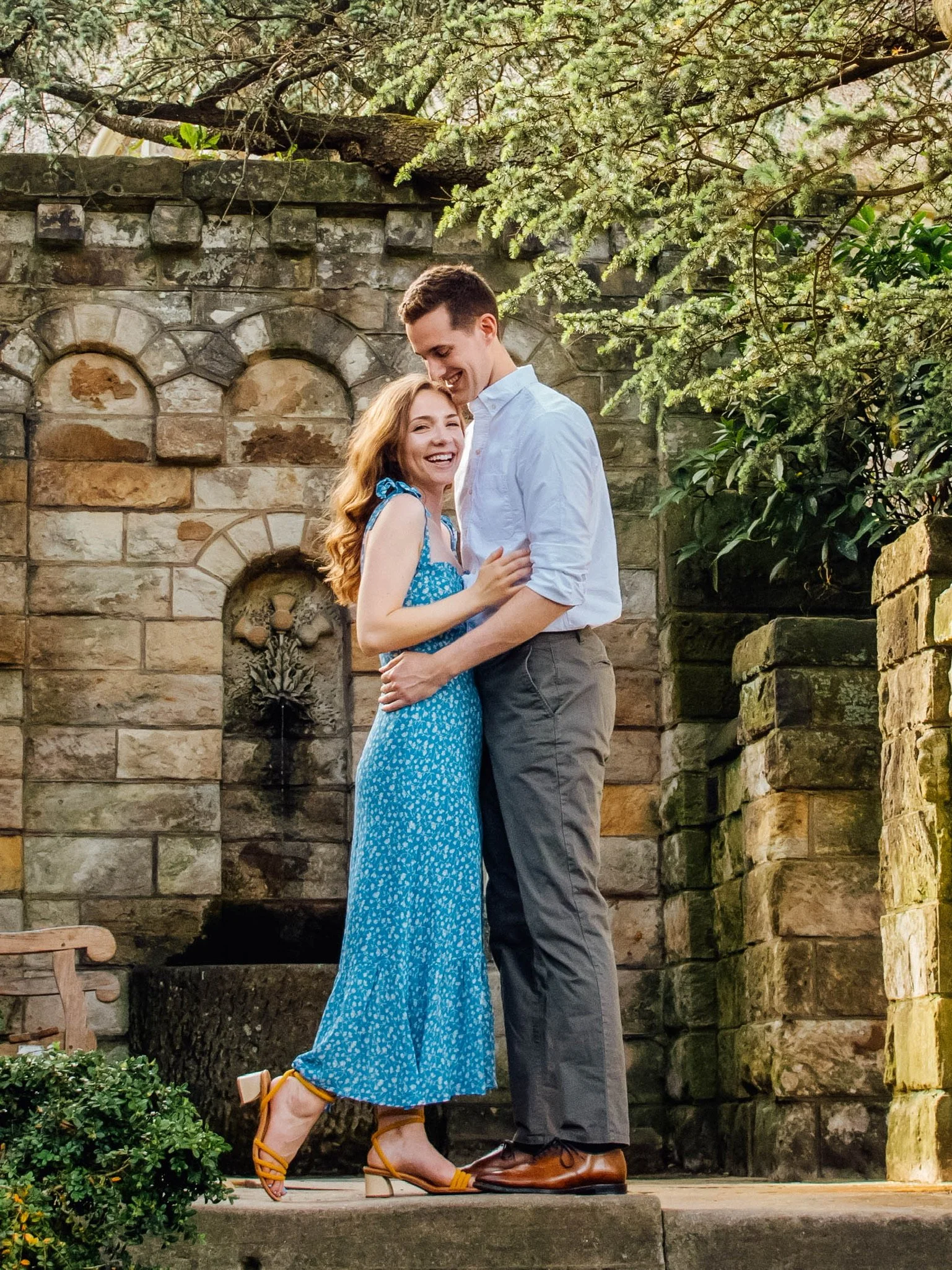 A young couple standing close together outdoors in front of a stone wall, smiling and looking at each other. The woman is wearing a blue dress with a small white pattern and yellow heels, and the man is wearing a white shirt and grey pants. The setti