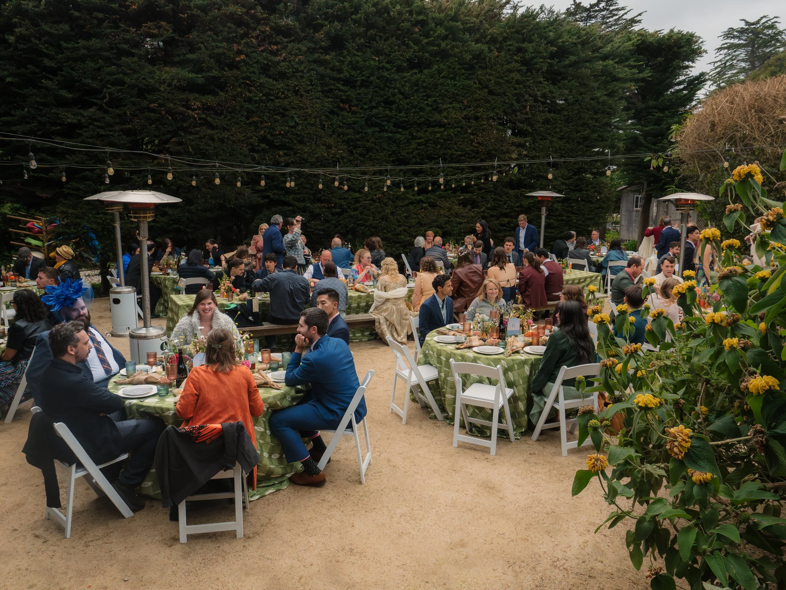 A large outdoor gathering with many people seated at decorated tables, some standing and socializing. The setting is surrounded by lush green trees and decorated with string lights and yellow flowers.
