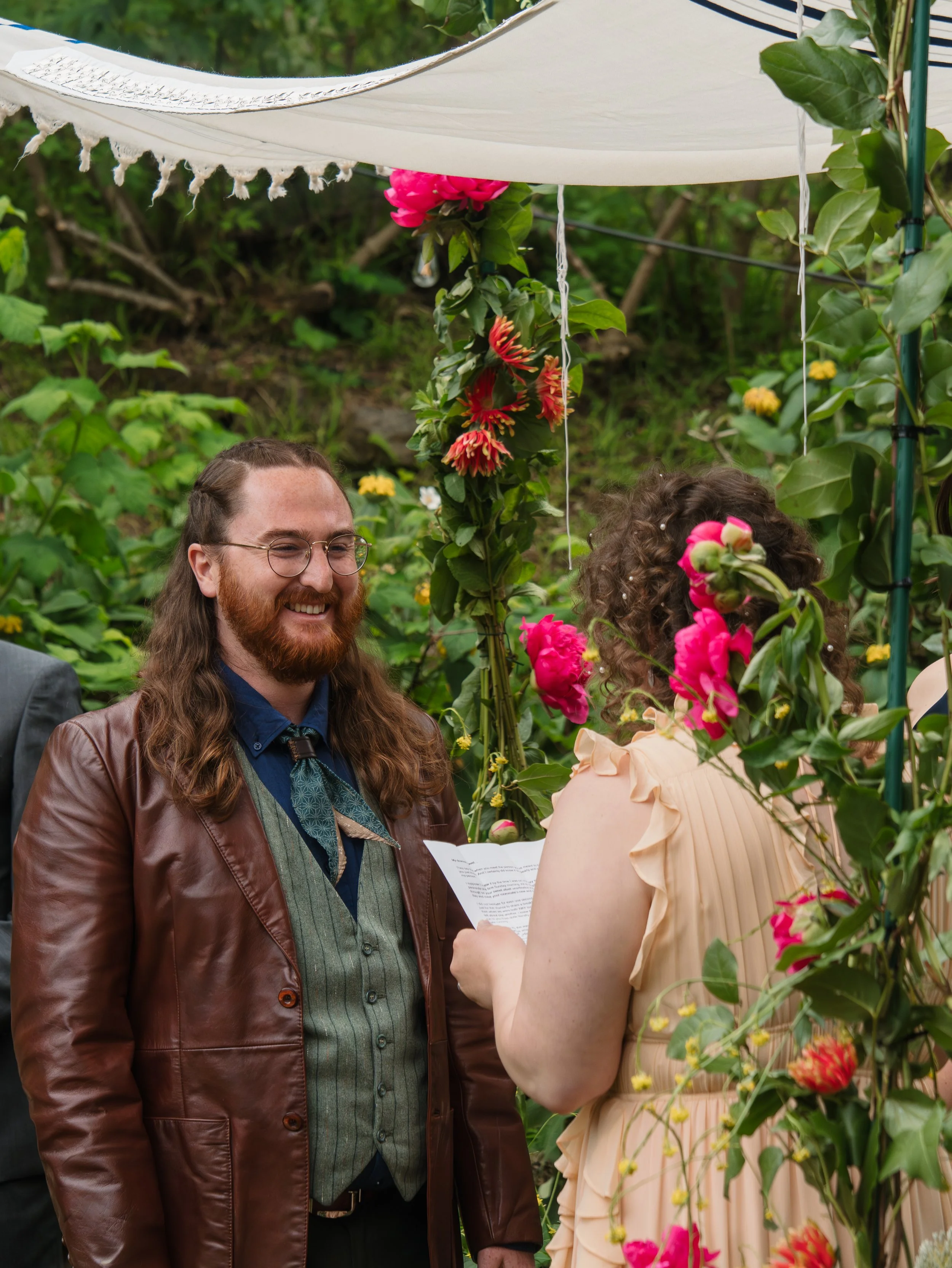 Man with long hair and glasses smiling during a wedding ceremony, looking at a woman with curly hair in a peach dress holding a paper, surrounded by plants and flowers.