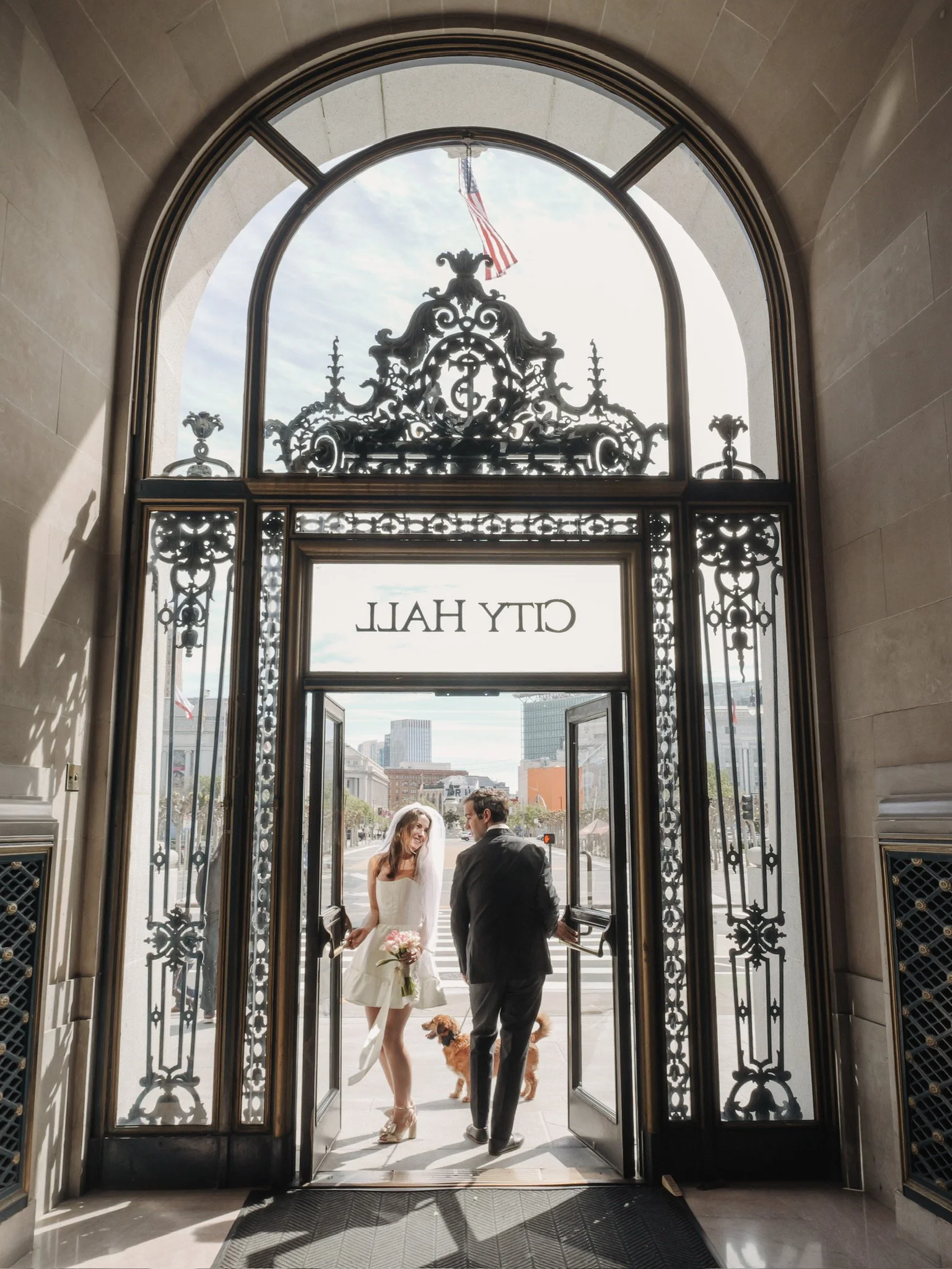 A bride and groom stand at the entrance of a building with an ornate wrought iron gate, holding hands and facing each other. The bride holds a bouquet of pink and white flowers, and both are accompanied by two dogs. The scene is backlit by daylight, 