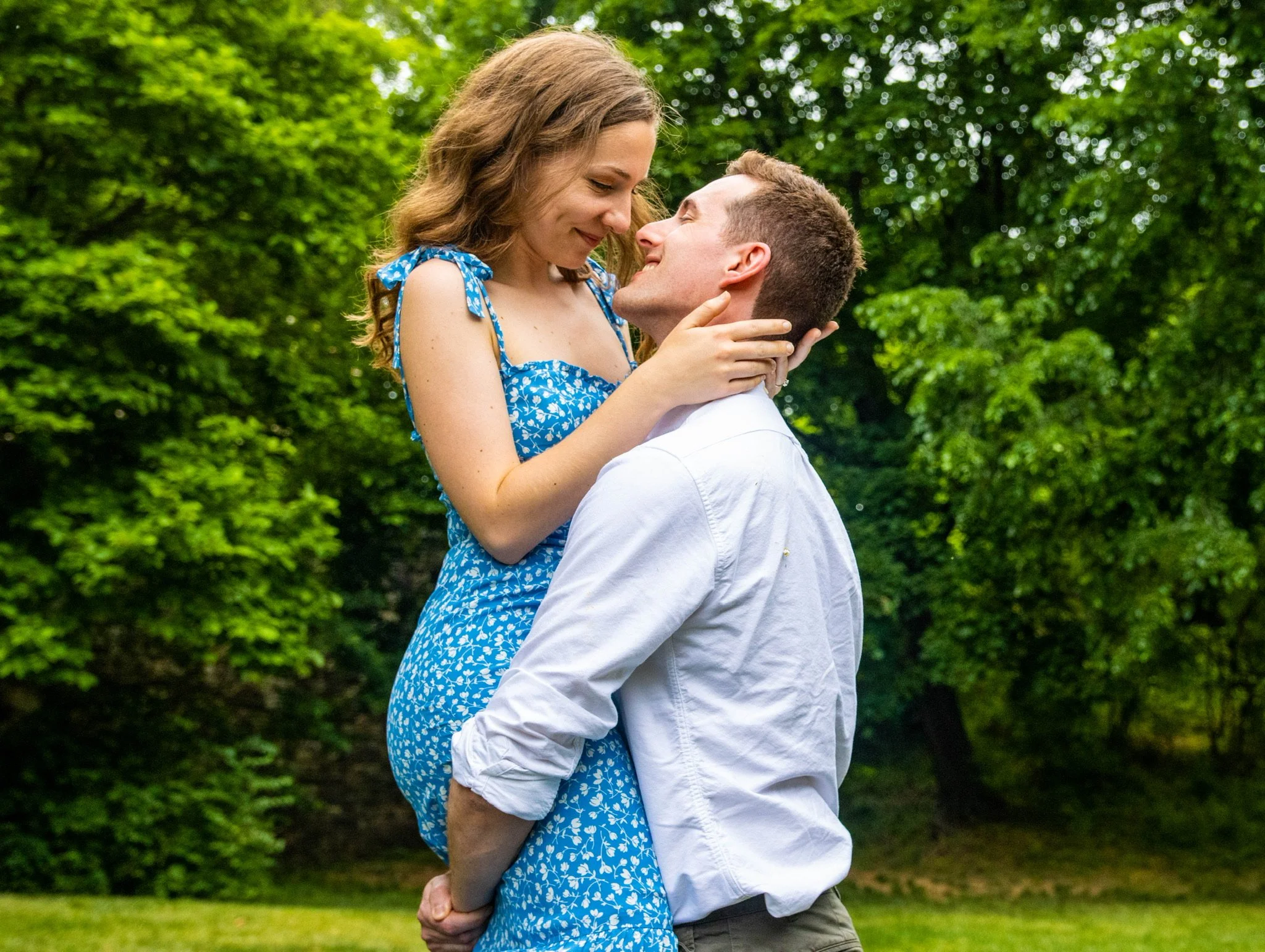A young man and woman are outdoors, smiling and close to each other, with green trees in the background.