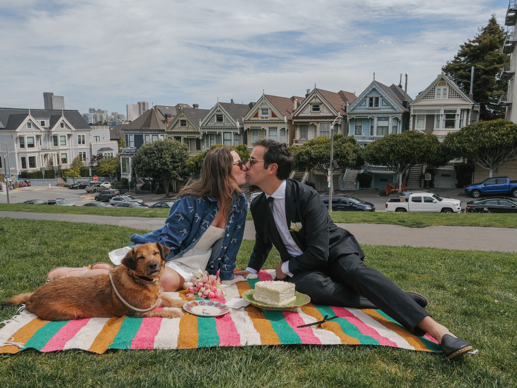 A couple kissing at a picnic on a colorful blanket, with a dog lying beside them and a cake with flowers nearby. There are houses and city buildings in the background.