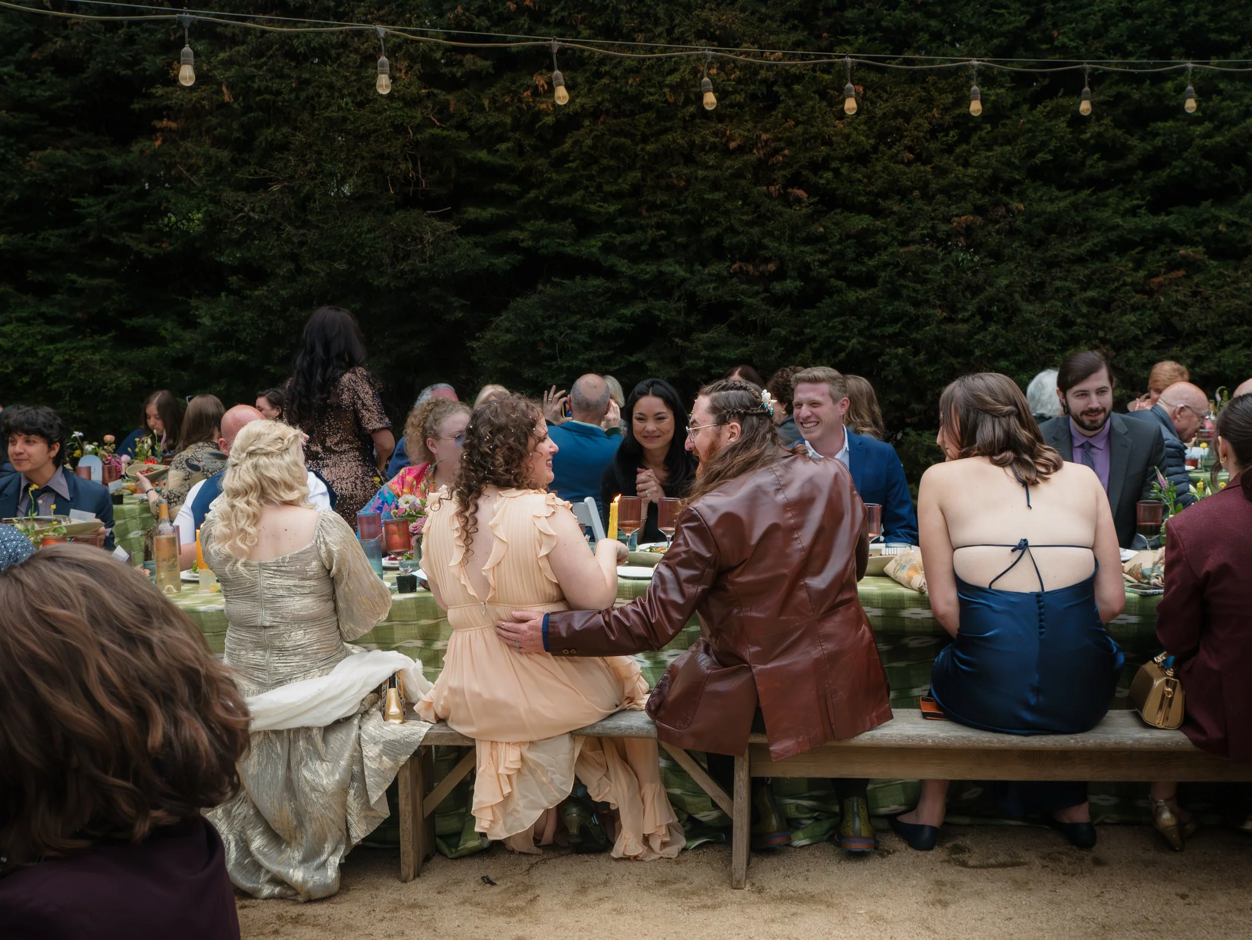 A group of people seated at long tables outdoors, enjoying the celebration, with string lights hanging overhead and greenery in the background.