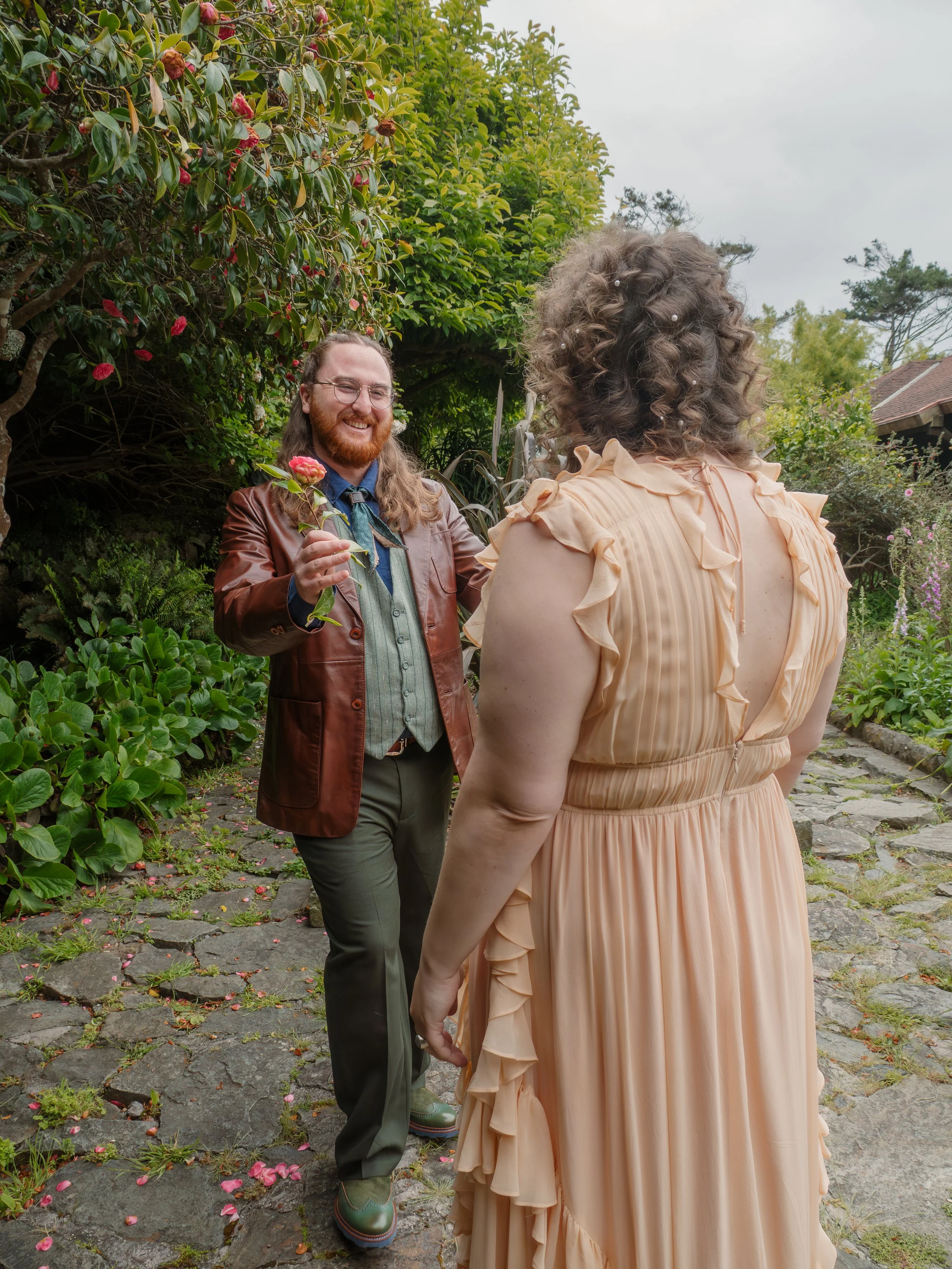 A man with long brown hair, a beard, glasses, wearing a brown leather jacket and a blue tie, is smiling and holding a pink flower. He is standing outside on a stone path, surrounded by green bushes and trees, talking to a woman with curly, shoulder-l