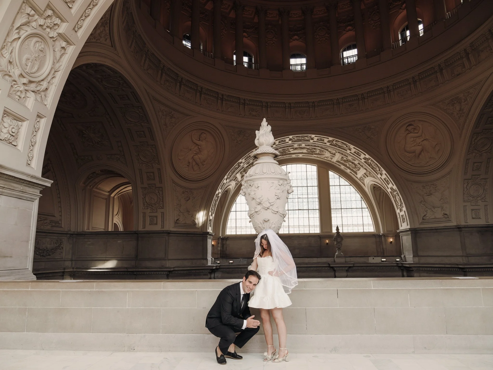 A couple posing at San Francisco City Hall with large windows and detailed architectural sculptures. The woman is wearing a white dress and veil, while the man is dressed in a black tuxedo. They are smiling and the man is kneeling on the ground.