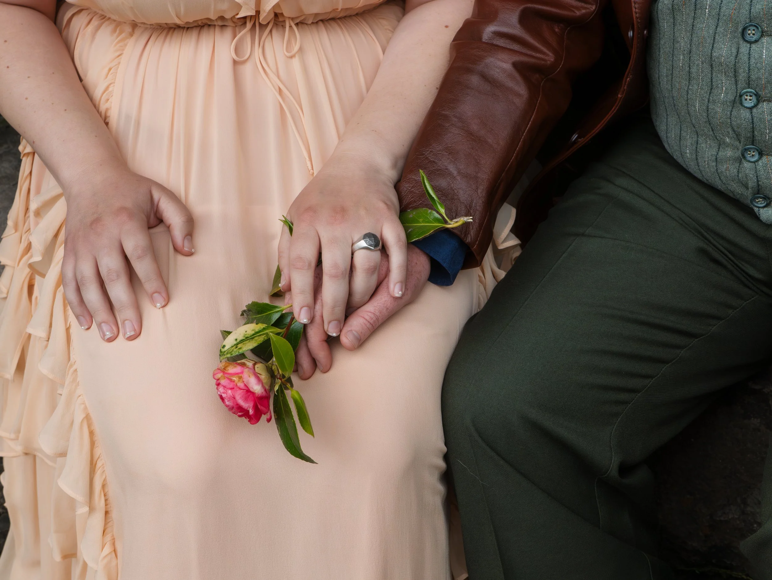 Close-up of two people holding hands, with one person's hand holding a small bouquet of pink flowers and green leaves, and the other person's hand resting on top. The people are wearing different clothing, one in a ruffled peach dress and the other i
