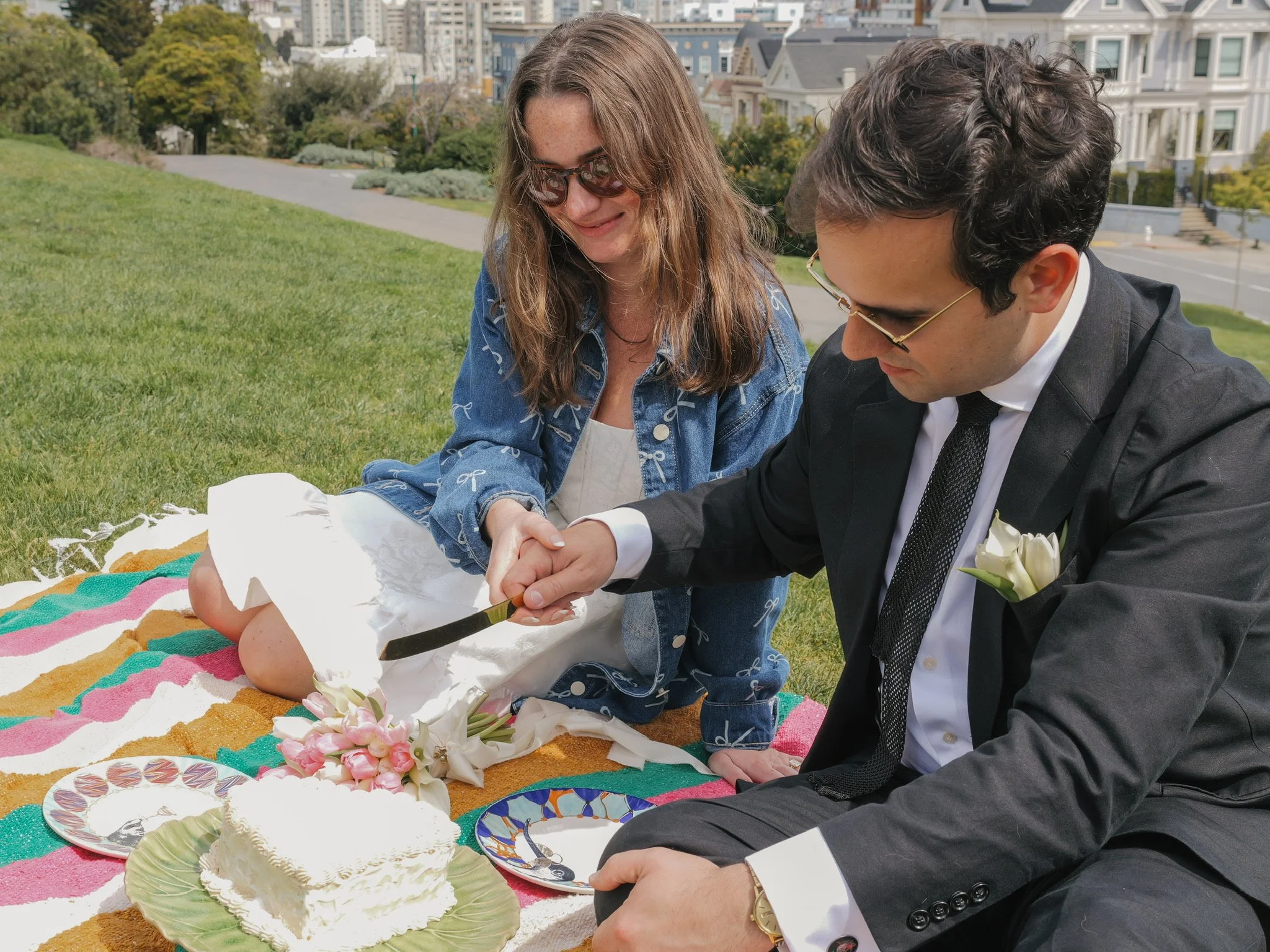 A couple, dressed in formal attire, is outdoors on a grassy area, sharing an intimate moment during a celebration. The woman is wearing sunglasses and a denim jacket, while the man is in a suit with a white shirt and tie. They are touching hands and 
