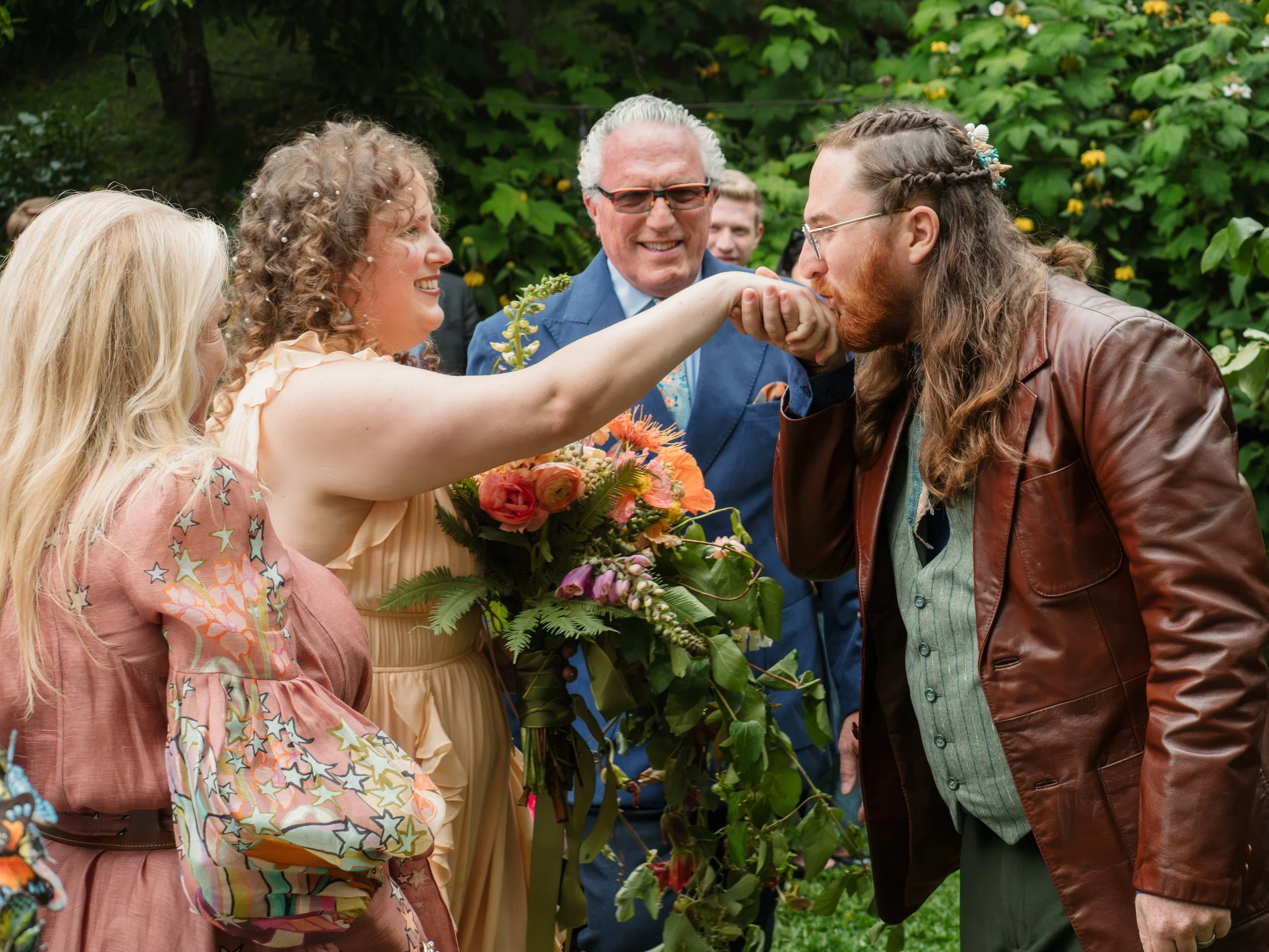 A man with long hair and glasses kissing a woman holding a bouquet of flowers during a wedding ceremony outdoors, with other guests and greenery in the background.