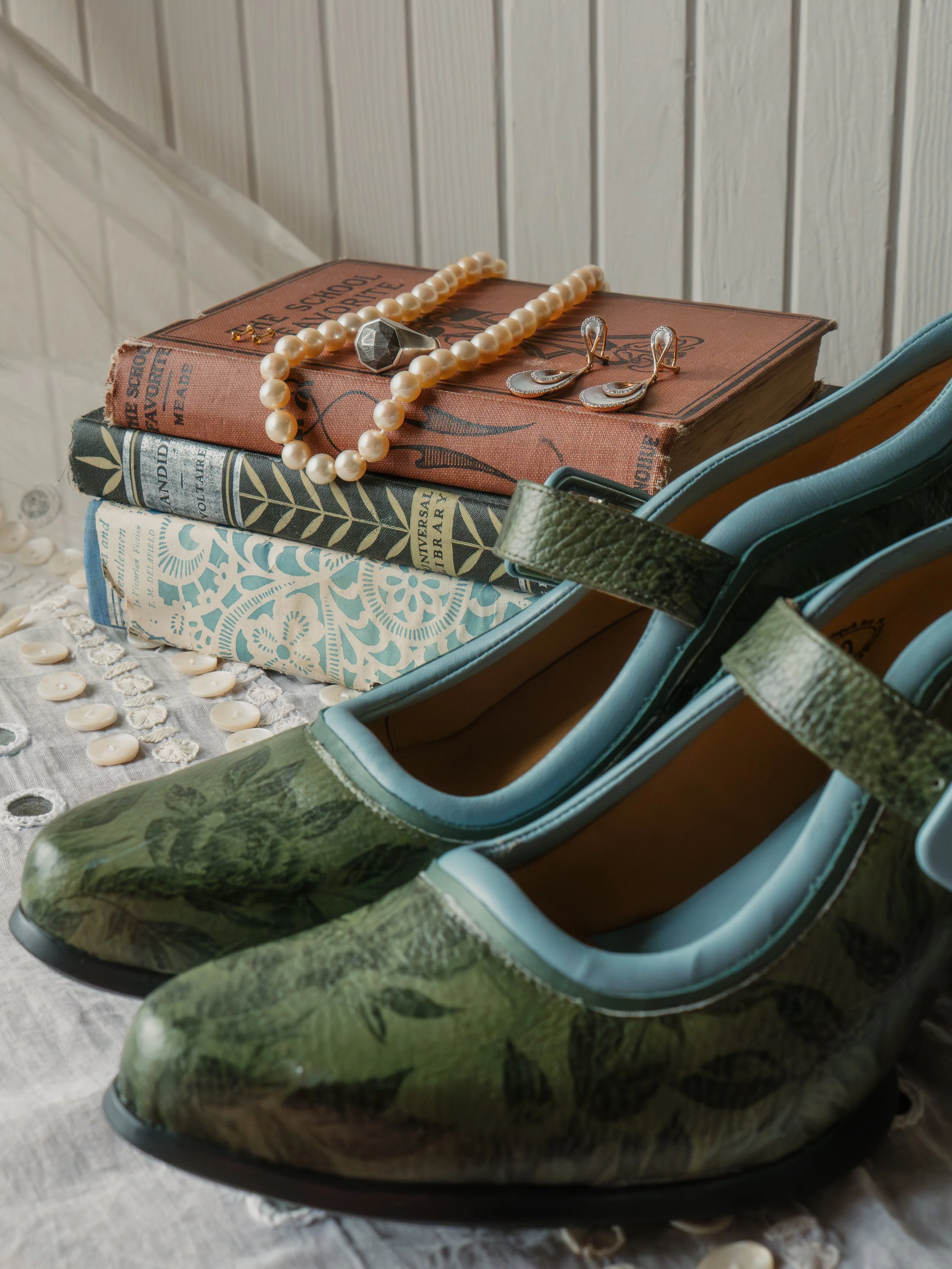 A pair of green heels, a stack of books, a pearl necklace, and earrings placed on top of the books.