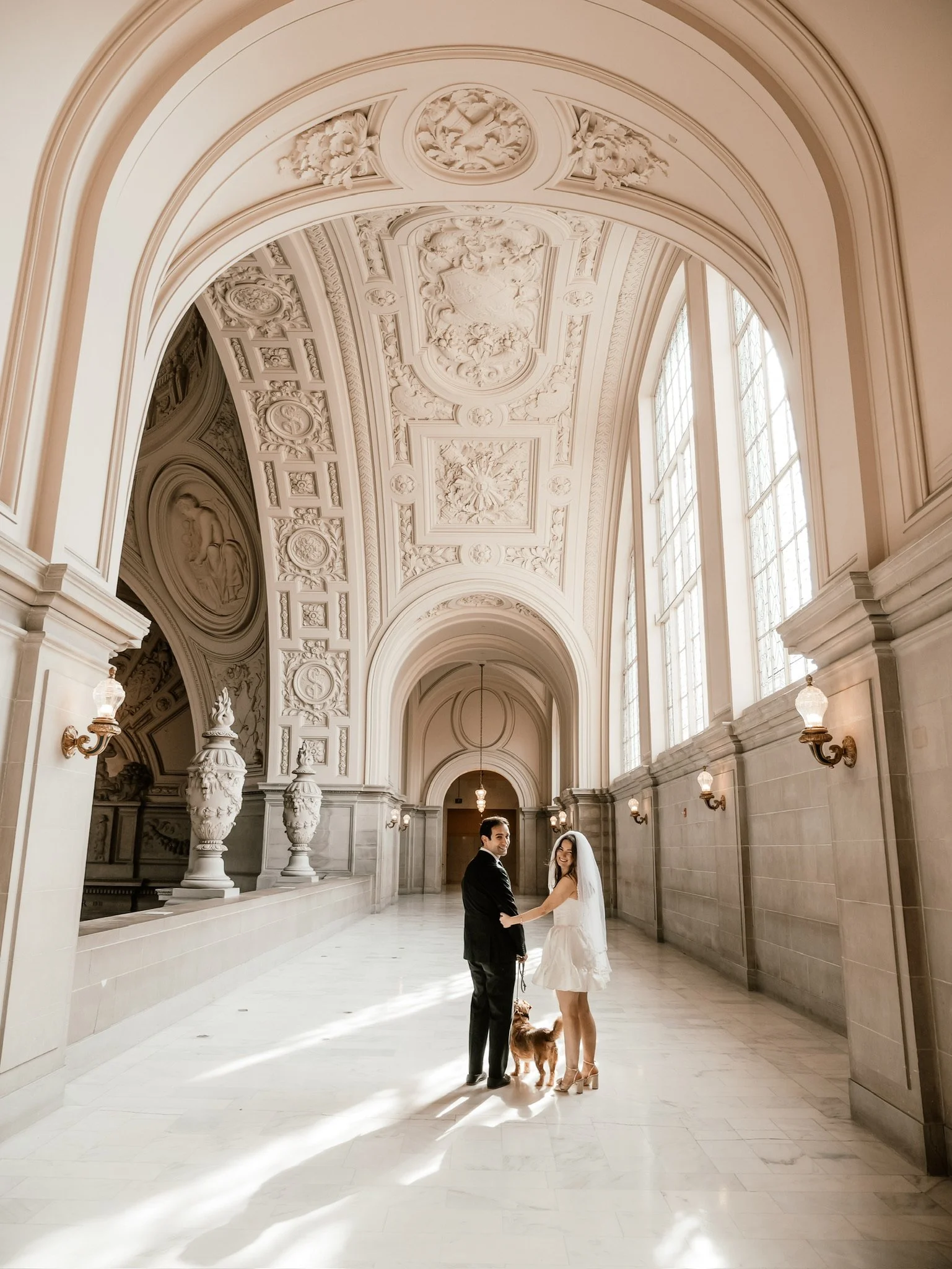 A newlywed couple with their dog at San Francisco City Hall with high ceilings, intricate moldings, large windows, and vintage wall sconces.