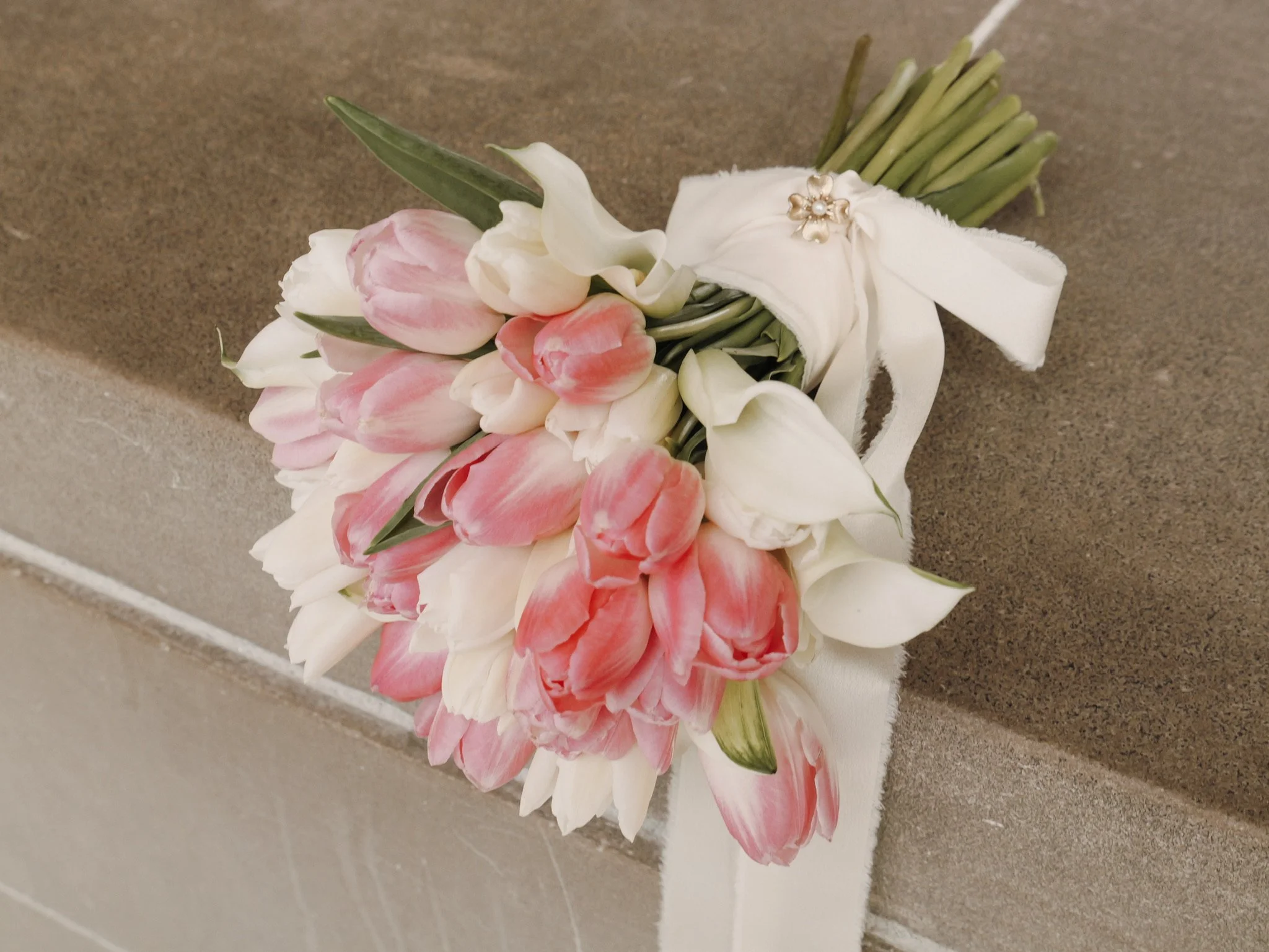 A bouquet of white and pink tulips with green leaves, decorated with a pearl and gold flower brooch, lying on a stone surface.