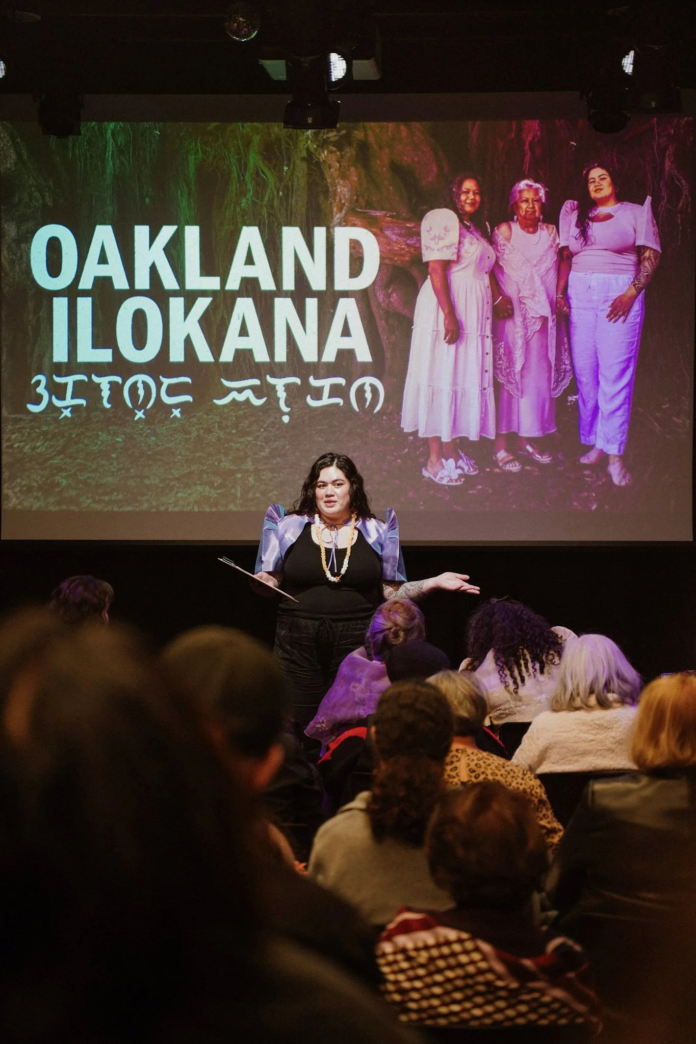 A woman wearing a black dress and patterned blue and purple jacket is speaking in front of a large screen with an image of three women dressed in white, standing in a natural setting. The screen has the words 'OAKLAND ILOKANA' and text in a different language.