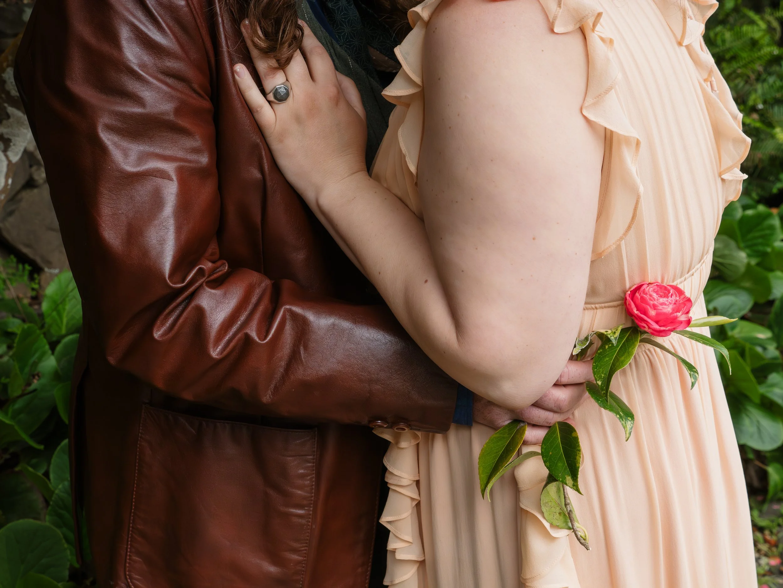 Close-up of a woman in a peach dress holding a pink flower and a man in a brown leather jacket with his arm around her, outdoors with green foliage in the background.