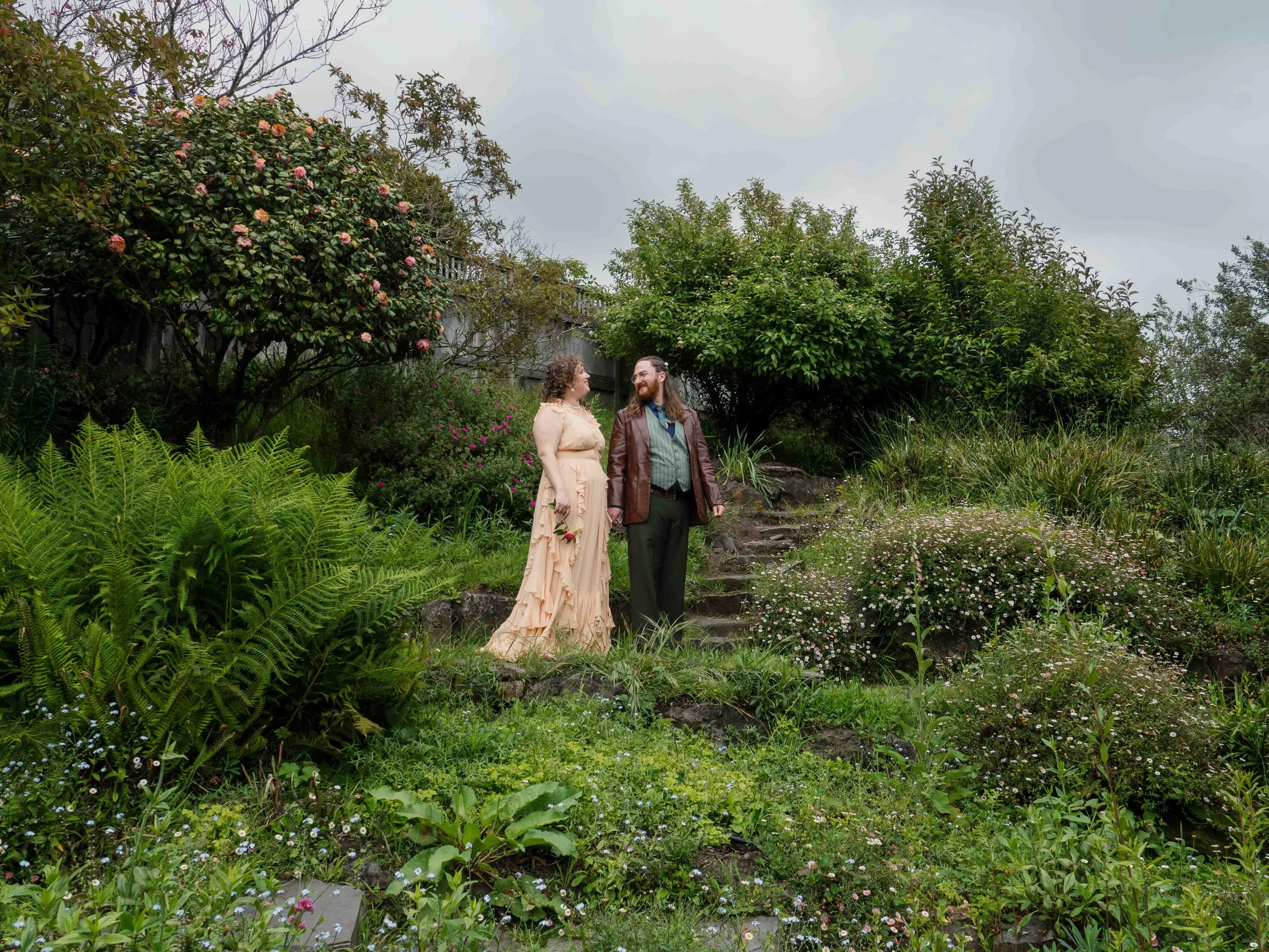 A couple standing together outdoors in a lush garden with various green plants and bushes, some flowering, on an overcast day.