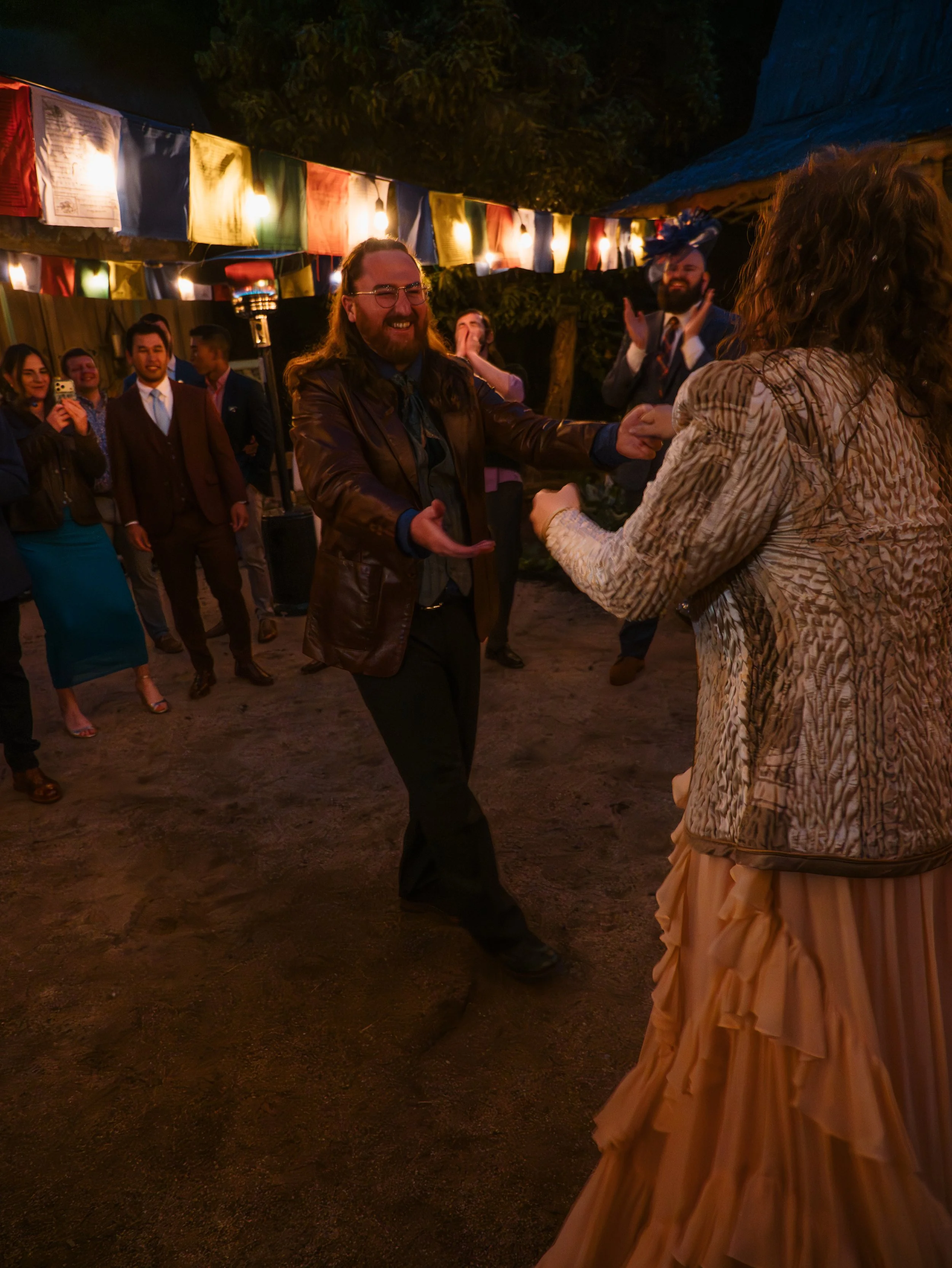 People dancing and celebrating at an outdoor event decorated with colorful flags and string lights at night.