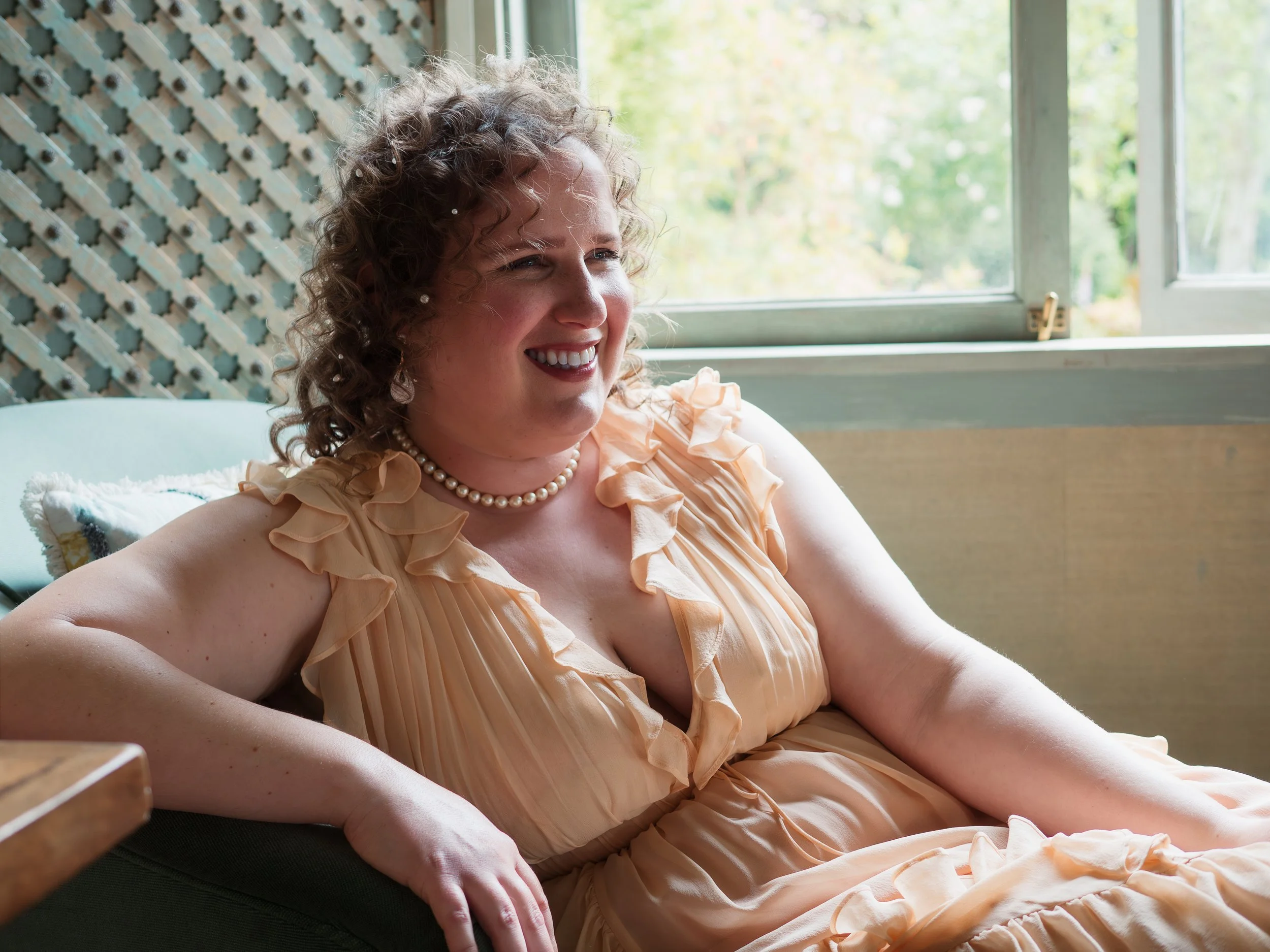 A woman with curly hair smiling and sitting on a couch by a window, wearing a peach-colored dress with ruffles and a pearl necklace.