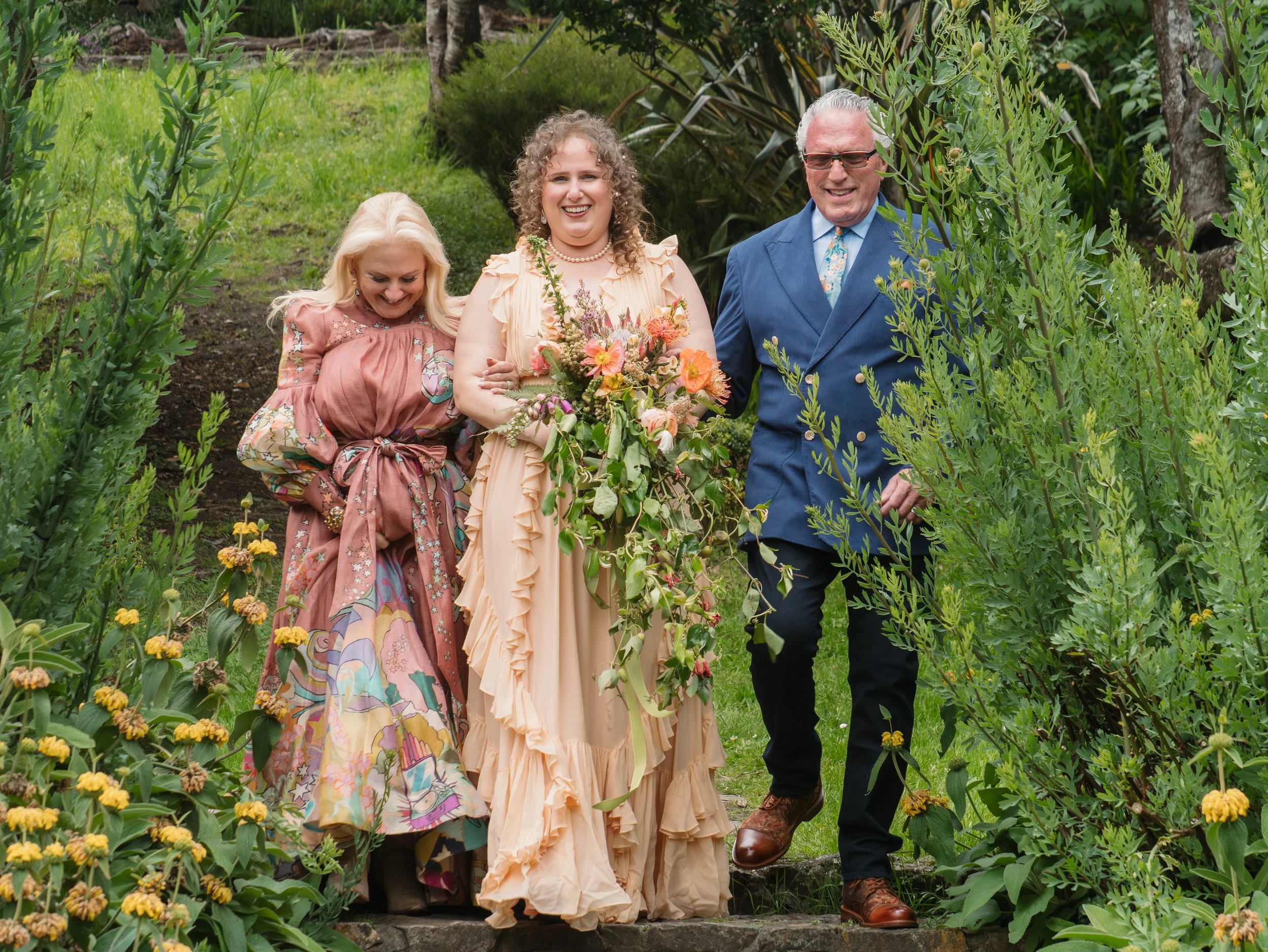 A group of three people, including a woman in a peach dress holding a bouquet of flowers, standing together on a garden path surrounded by greenery and yellow flowers, smiling and enjoying a sunny day.