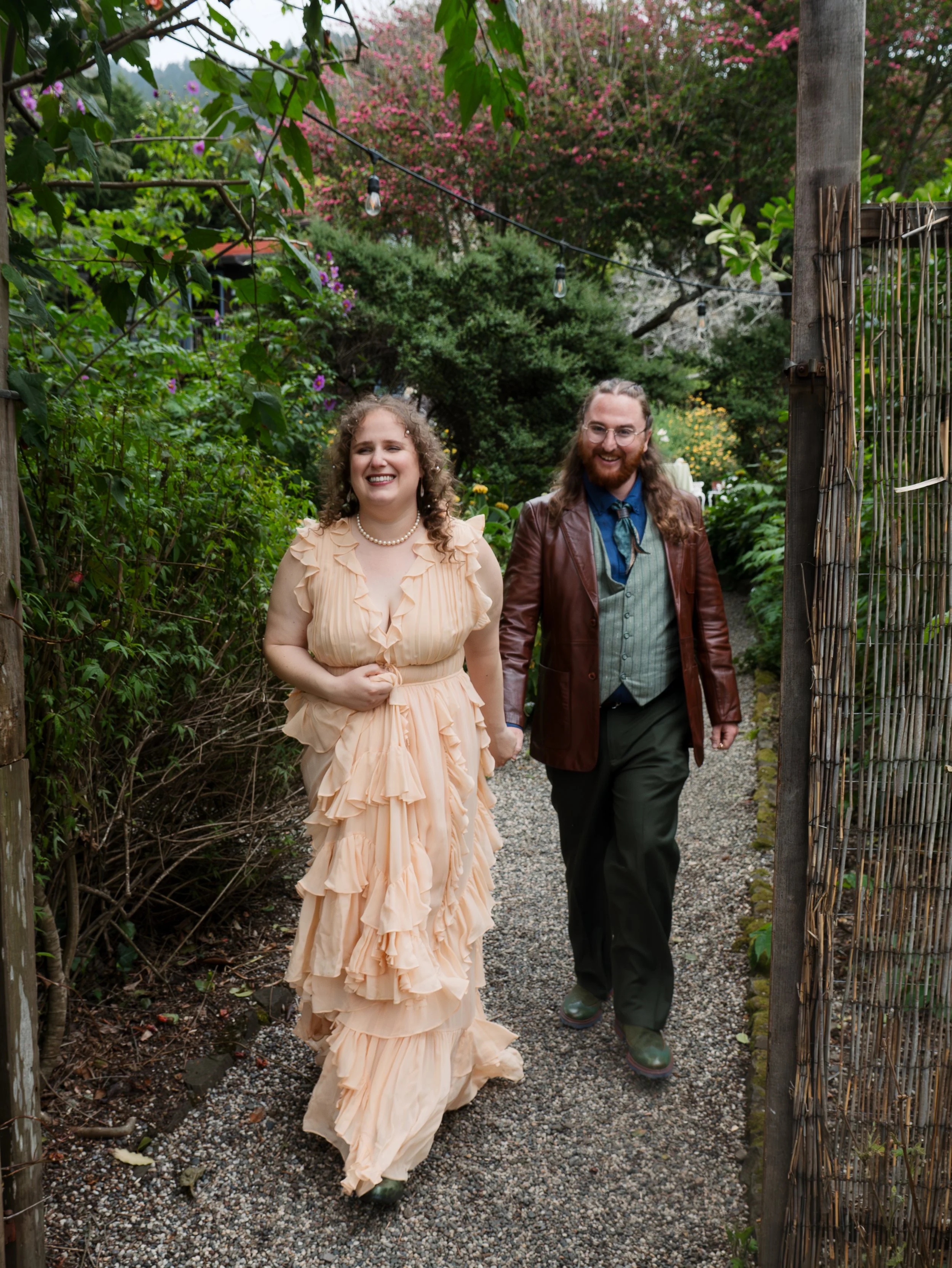 A woman in a peach ruffled dress and pearl necklace walking with a man in a brown leather jacket, green shirt, and dark pants on a garden path surrounded by lush greenery and colorful flowers.