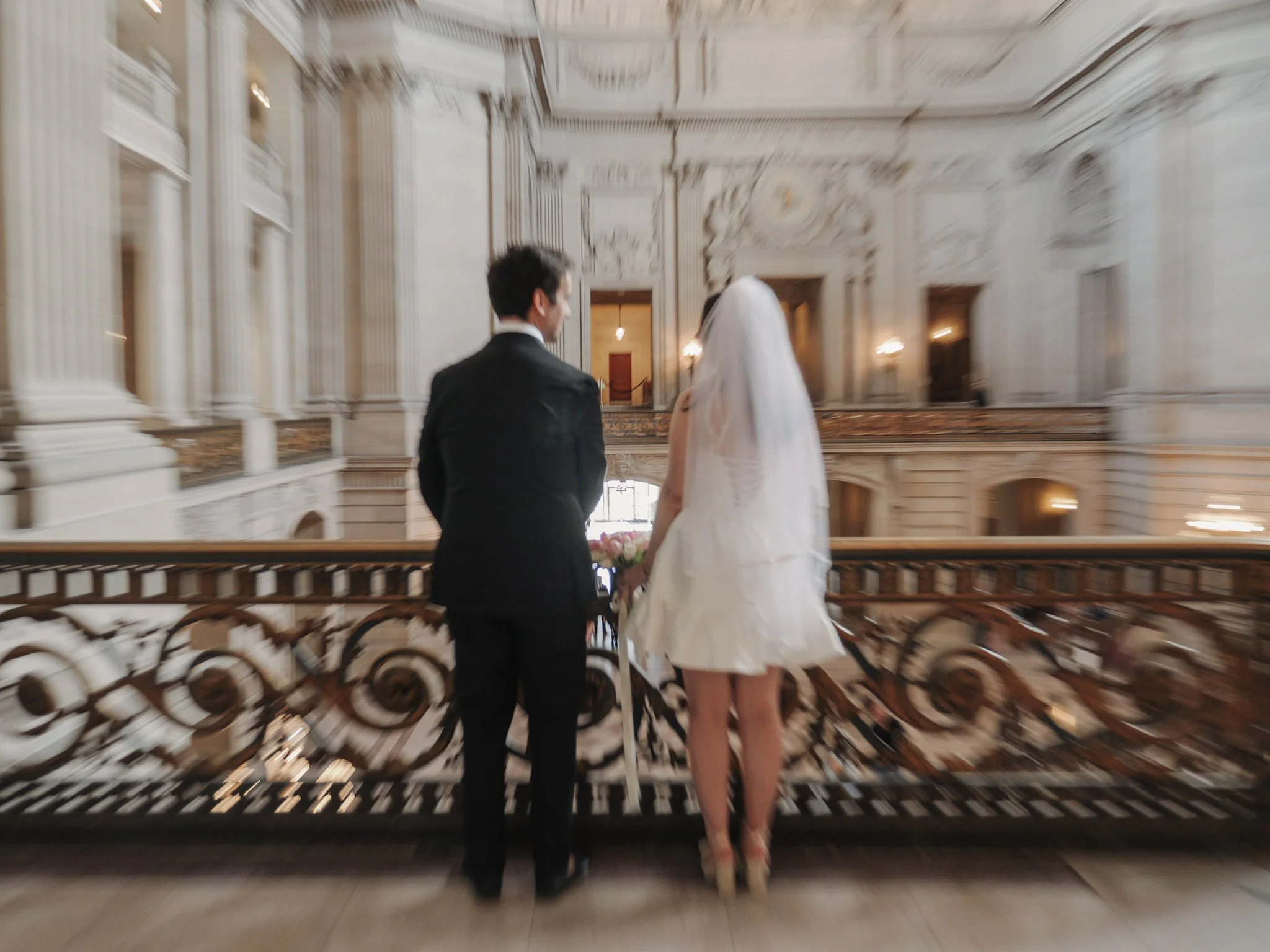 A bride and groom holding hands and walking up an ornate staircase in San Francisco City Hall with elaborate architectural details.