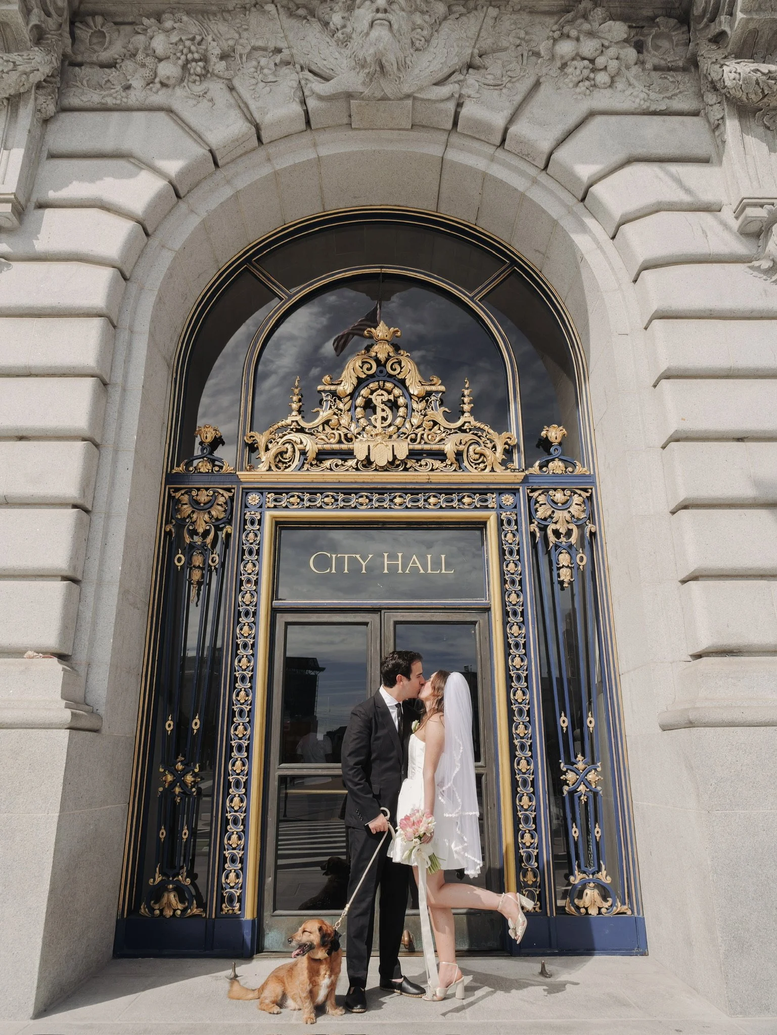 A wedding couple kissing in front of San Francisco City Hall entrance, with a dog at their feet and a reflection of the city in the glass door.