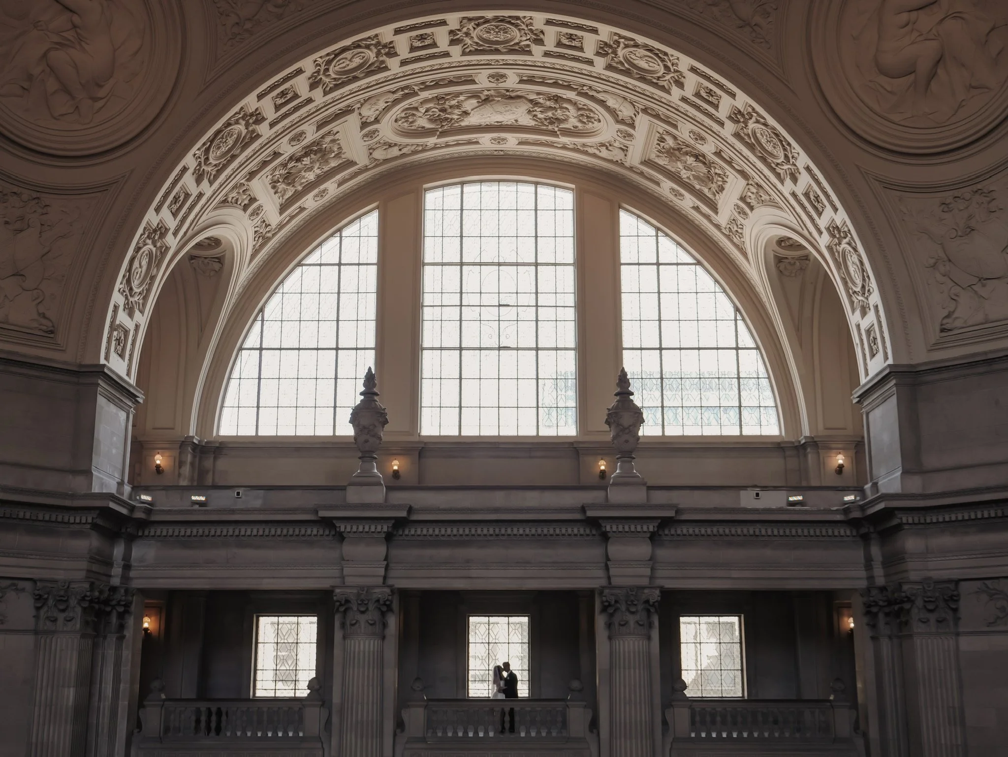 The image shows San Francisco City Hall with large arched windows, intricate decorative moldings, and two people standing close together in front of the windows, silhouetted against the light.