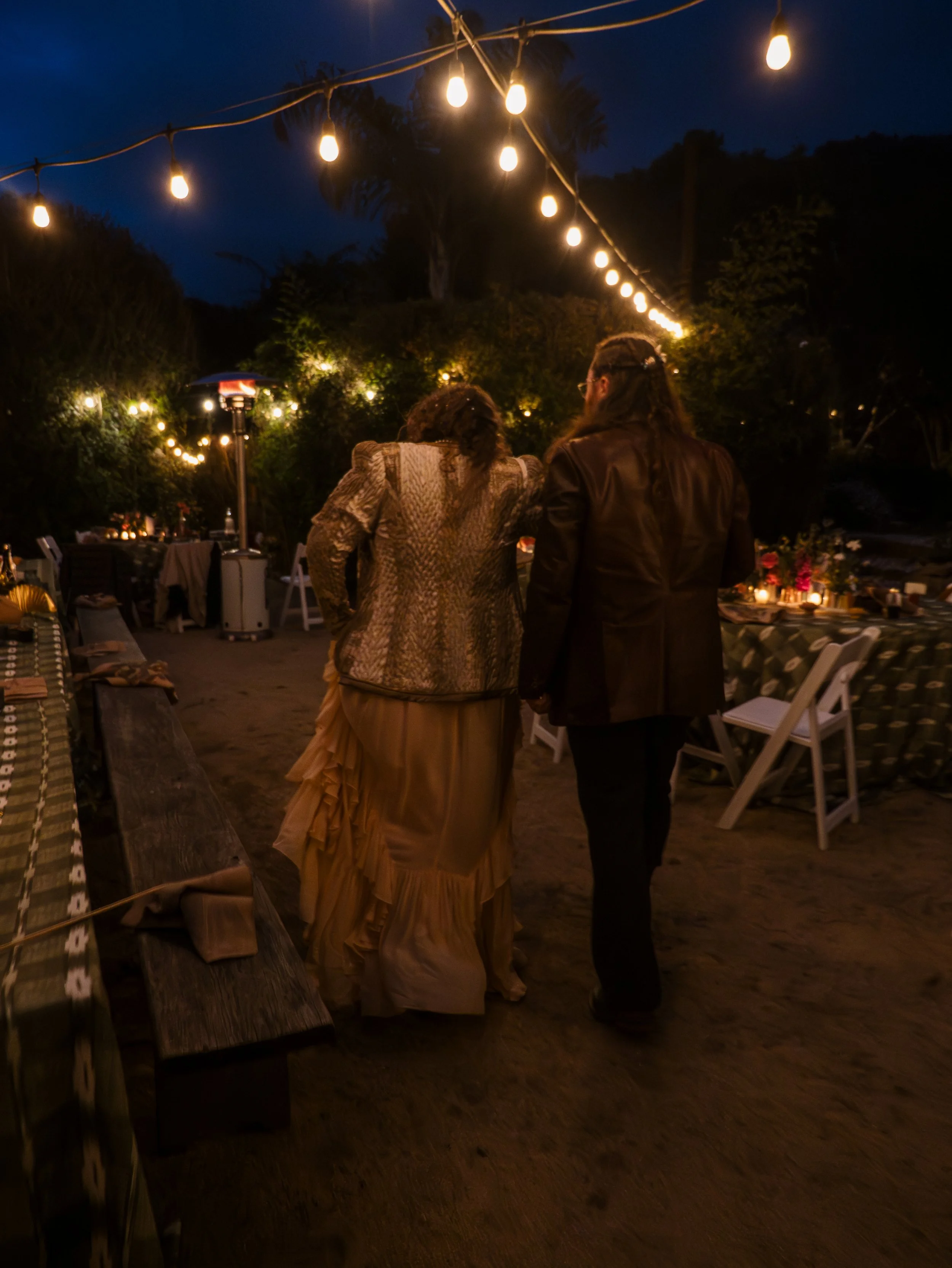 People standing outdoors at night under string lights, with tables and chairs around, trees in the background, and a patio heater.